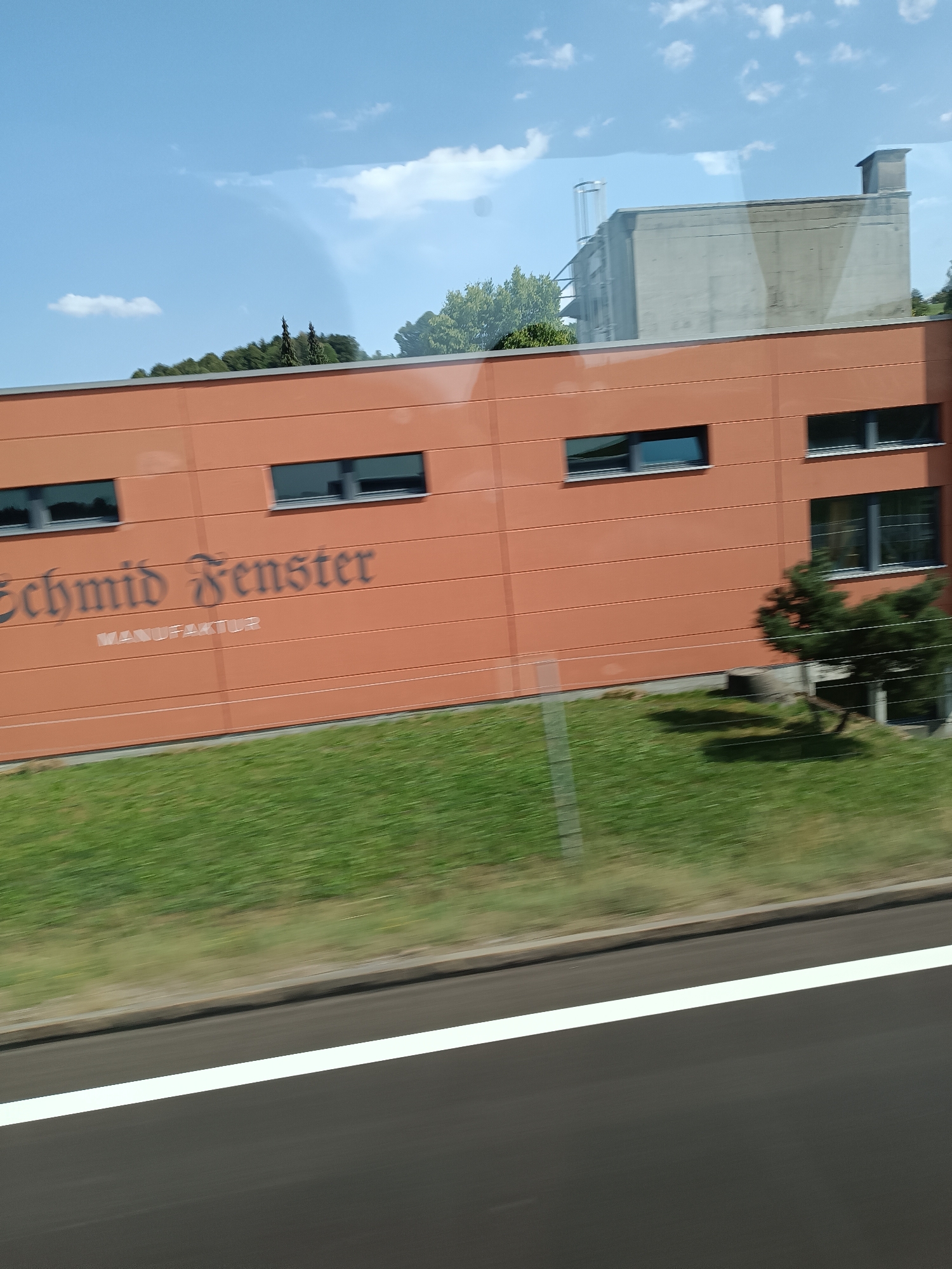 This image shows a side view of a modern, orange-colored building with dark-framed windows and the text "Schmid Fenster MANUFAKTUR" on its facade. The building appears to be a factory or workshop, possibly for manufacturing windows. In front of the building, there is a strip of green grass and a small, sparse tree. The background includes a bright blue sky with scattered white clouds, and a glimpse of other buildings and trees further in the distance. The foreground shows a dark asphalt road with a prominent white line, suggesting the photo was taken from a moving vehicle on a highway or similar road. The overall scene suggests a sunny day in an industrial or semi-industrial area.