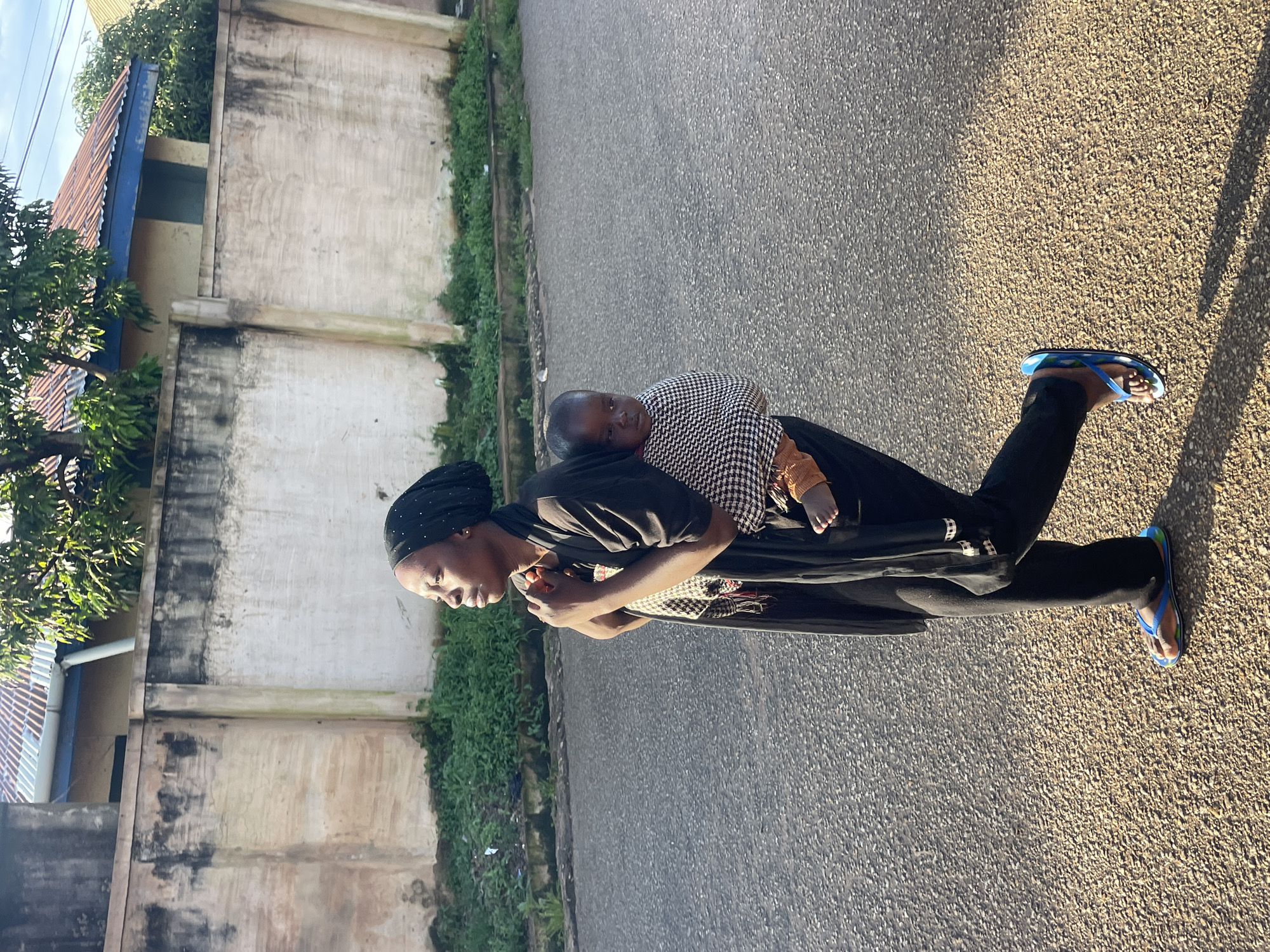 A woman wearing a black hijab and black clothing walks along a paved road in Jos, Nigeria. She is carrying a baby on her back, wrapped in a black and white patterned cloth. The baby is wearing orange socks. The woman is wearing blue flip-flops. A concrete wall, partially covered in moss, runs along the side of the road, with green vegetation growing at its base. Behind the wall, a building with a corrugated metal roof and blue trim is visible. Trees with green foliage are also present. The sun is casting shadows on the road.