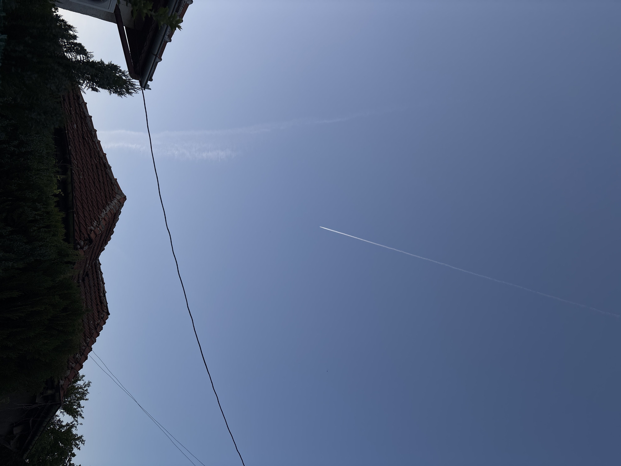 The image shows a blue sky with two contrails, taken from the city of Lom, Bulgaria. The lower left corner of the image includes a building with a red tile roof, covered in greenery, and a black power line.