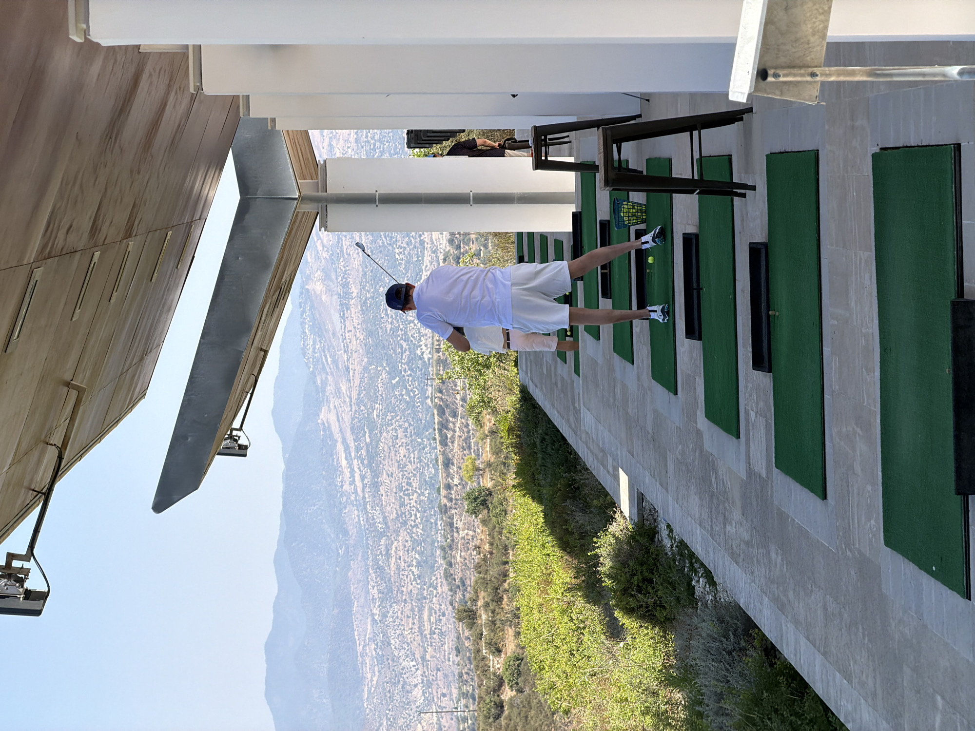 In Tsada, Cyprus, at a golf driving range, a person wearing a white polo shirt, white shorts, and a blue baseball cap stands on a green mat, mid-swing with a golf club. To their left, another person in pink shorts and a white top is visible. The driving range is characterized by a tiled walkway, multiple green practice mats, and a basket of golf balls. White pillars support a large, angled wooden roof structure. In the background, a vast, dry, mountainous landscape stretches under a clear blue sky. The scene is illuminated by bright, natural daylight.