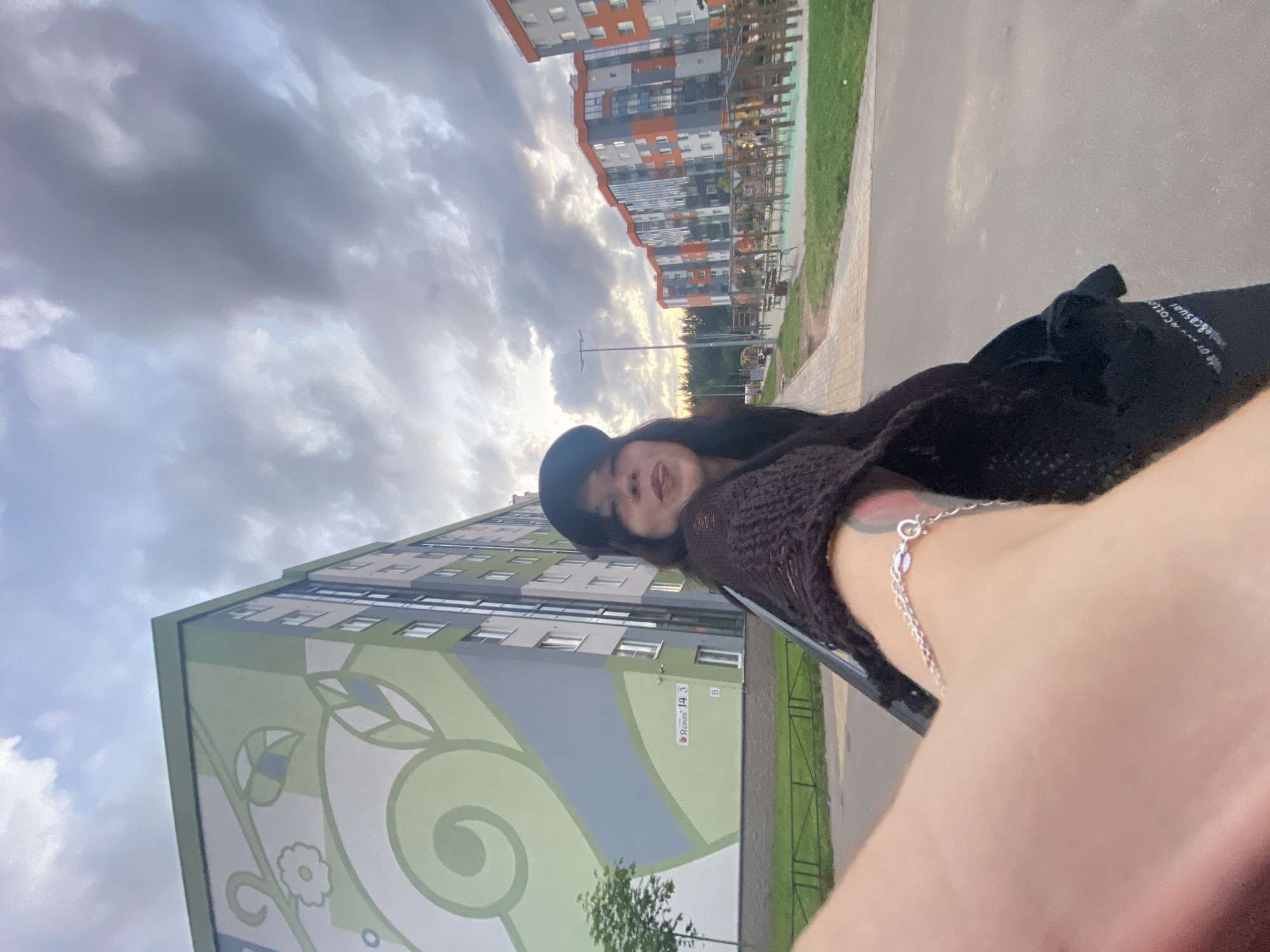 A first-person perspective, selfie-style photograph taken outdoors in the city of Yanino-1, Russia, captures a young woman with dark hair, wearing a black baseball cap and a dark, open-knit sweater. She is extending her right arm towards the camera, with her hand and part of her forearm visible, adorned with a silver bracelet. Her head is turned slightly, and she is sticking her tongue out playfully.
Behind her, on the left, is a multi-story residential building with a large, stylized green mural featuring floral and leaf motifs. A street sign on the building reads "Ясная 14, 3". To the right of this building, further in the background, are several other modern residential buildings with orange and gray facades, and an outdoor playground area with wooden structures and green grass. The sky above is overcast with dramatic, grey clouds. The overall setting appears to be a residential urban environment with a playground and well-maintained buildings.