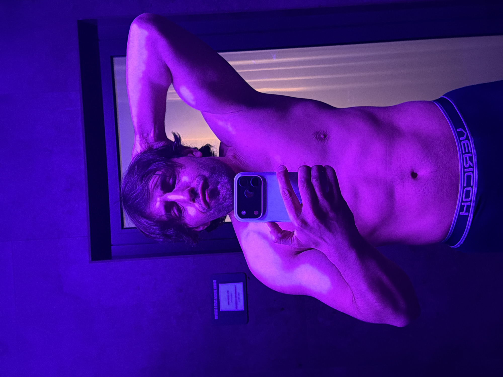 A shirtless man is taking a selfie in a mirror. He is wearing underwear with the brand name "EBICC" on the waistband. The environment is dimly lit with a purple hue. There is a window behind him with light coming through. There is a sign on the wall below the mirror. The image does not provide enough information to determine the location in the city of Krasnoyarsk, Russia.