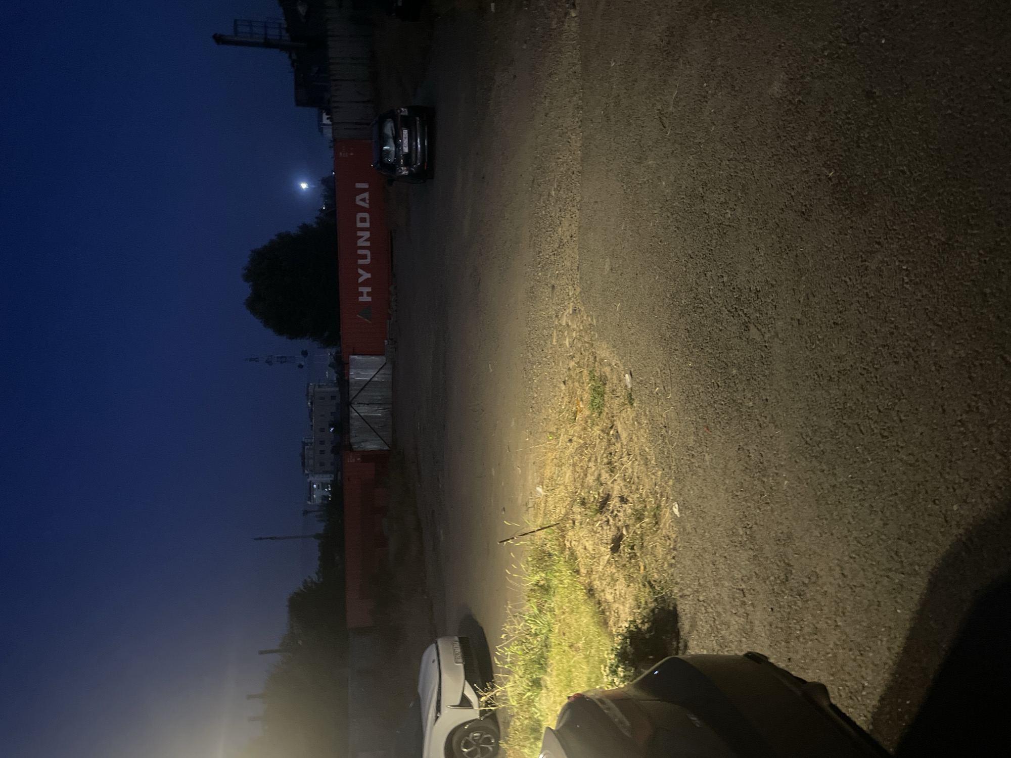 The image depicts an outdoor scene at dusk or night in Dushanbe, Tajikistan. In the background, a large, orange shipping container with the word "HYUNDAI" printed in white letters is visible. Behind the container, a few buildings and industrial structures are dimly lit against a dark blue sky. The foreground shows a paved area, likely a parking lot or street, with a patch of dry grass and weeds on the left. Two cars are present: a white car is parked on the left, and a dark-colored SUV is parked further back on the right. The headlights of the white car are illuminated, casting a bright light onto the grassy area. The overall impression is one of an industrial or commercial area at the end of the day.