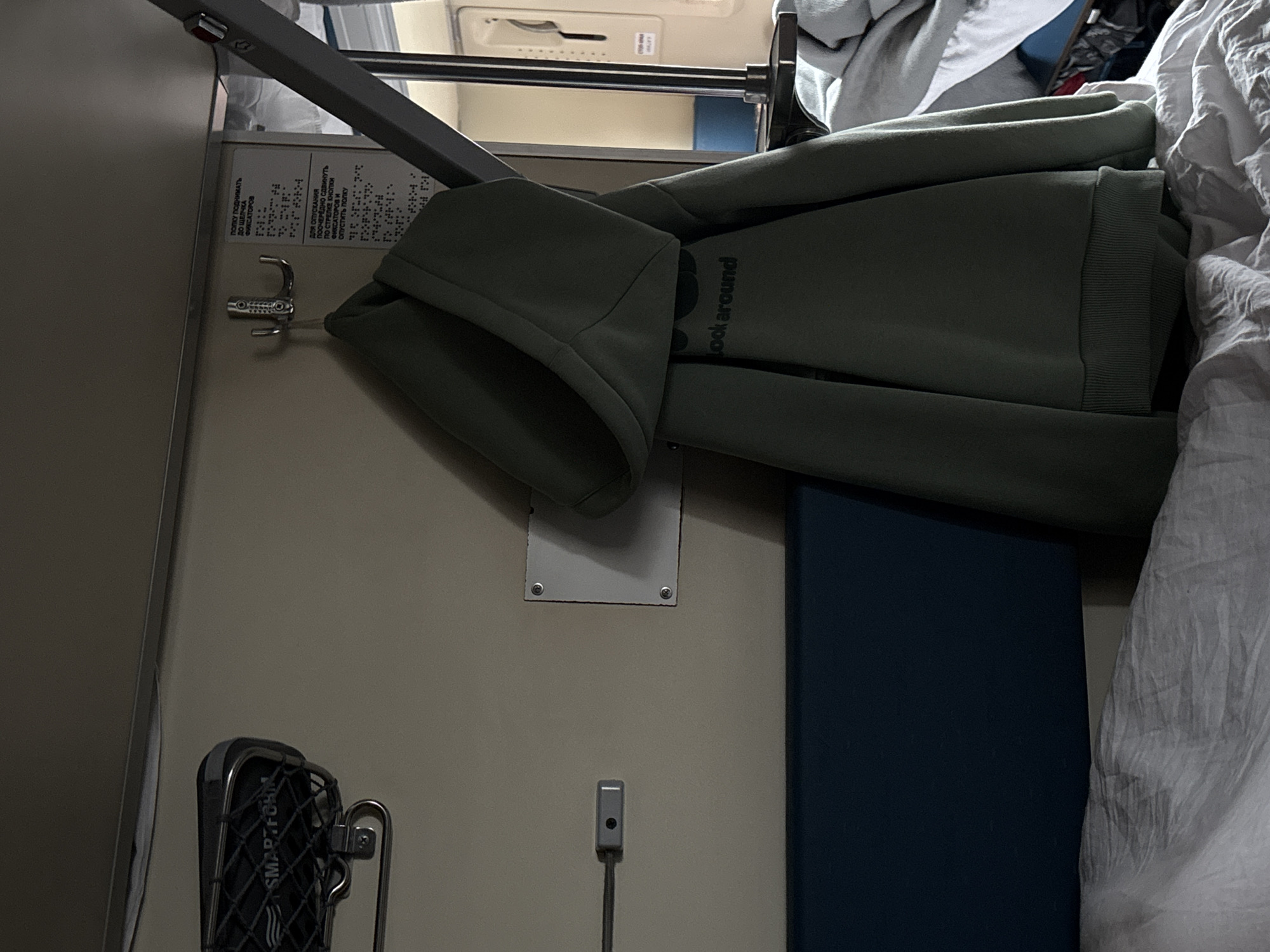 The image displays the interior of a train compartment, located within a carriage associated with Kargat, Russia. The scene is static and dimly lit. On the upper left, a dark-colored overhead luggage rack with a mesh base, marked "SMARTFORM," is affixed to a cream-colored wall. Below it, a small, horizontal rectangular fixture is mounted. Centrally positioned, a triple silver-toned metal hook suspends a dark green hooded sweatshirt, partially revealing the text "look around" on its front. Adjacent to the hook on the right, a white laminated sign displays instructions in Russian text and Braille regarding the operation of a shelf or bunk, specifying how to raise it until fixators click and how to lower it. Below the sign, a white square panel secured by four screws is visible. The lower section of the image features a dark blue upholstered element, possibly a backrest, over which rumpled white bedding lies on a bunk. To the far right, a vertical metallic bar and a glimpse of a door or window frame are present, revealing another blue surface beyond. No individuals are present in the scene.