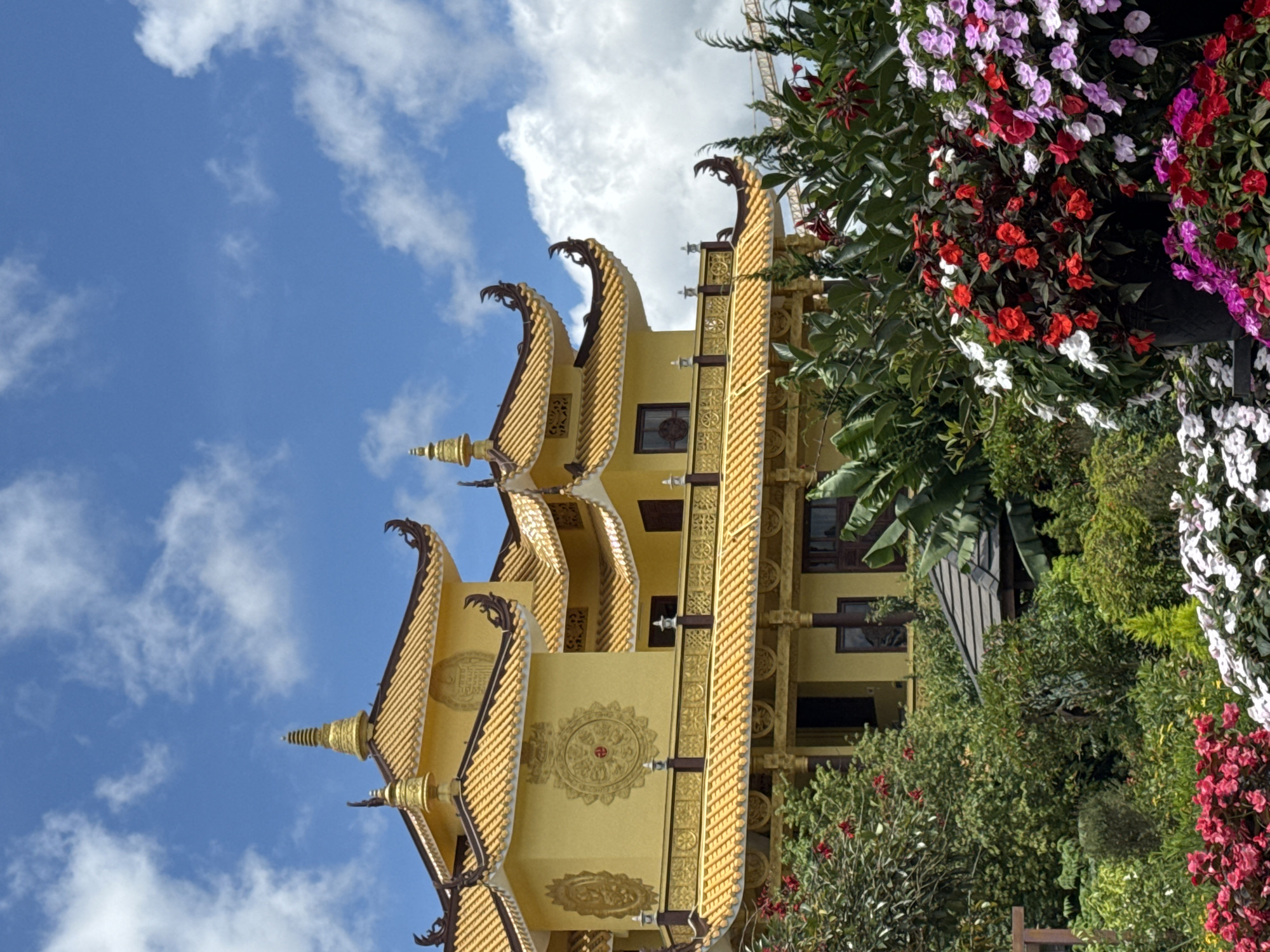 A bright yellow, multi-tiered Buddhist temple with ornate, upturned eaves and golden decorative elements stands against a bright blue sky with scattered white clouds. The temple is surrounded by lush greenery, including trees and vibrant flowers in shades of red, pink, white, and purple. The overall impression is one of serene beauty and traditional architecture. This is likely the Linh Phuoc Pagoda in Da Lat, Vietnam, a notable Buddhist temple known for its intricate mosaics and distinctive architecture.
