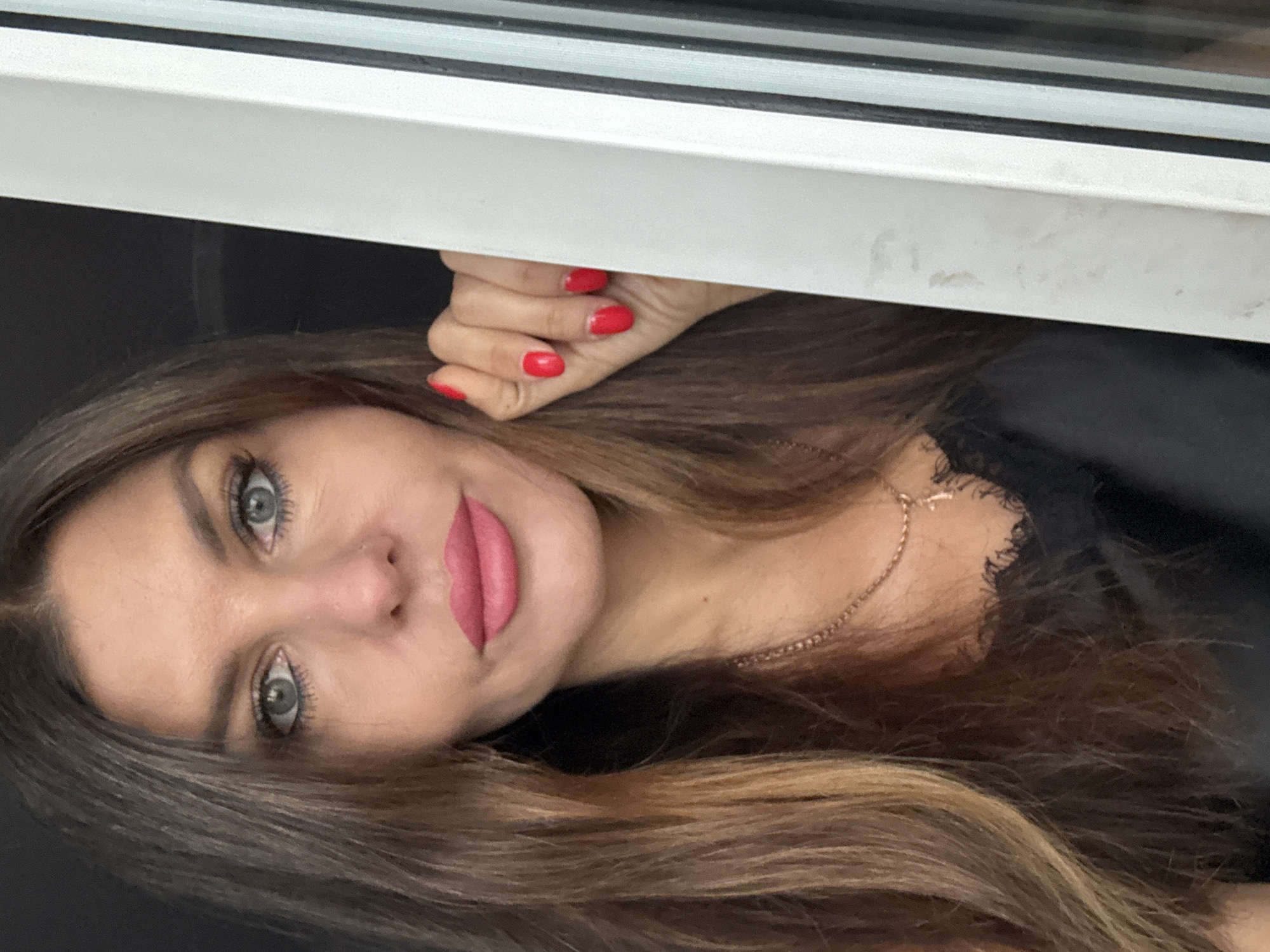 A close-up selfie shows a fair-skinned woman with long, wavy brown hair, blue eyes, and pink lipstick, looking directly at the camera. Her right hand, with bright red nail polish, is raised to her face, resting on the edge of what appears to be a window frame. She is wearing a black top with lace detailing and a gold necklace with a cross pendant. The background is dark, with the right side of the image showing a white frame with black vertical blinds or shutters. The location is the city of Chaikovsky, Russia.