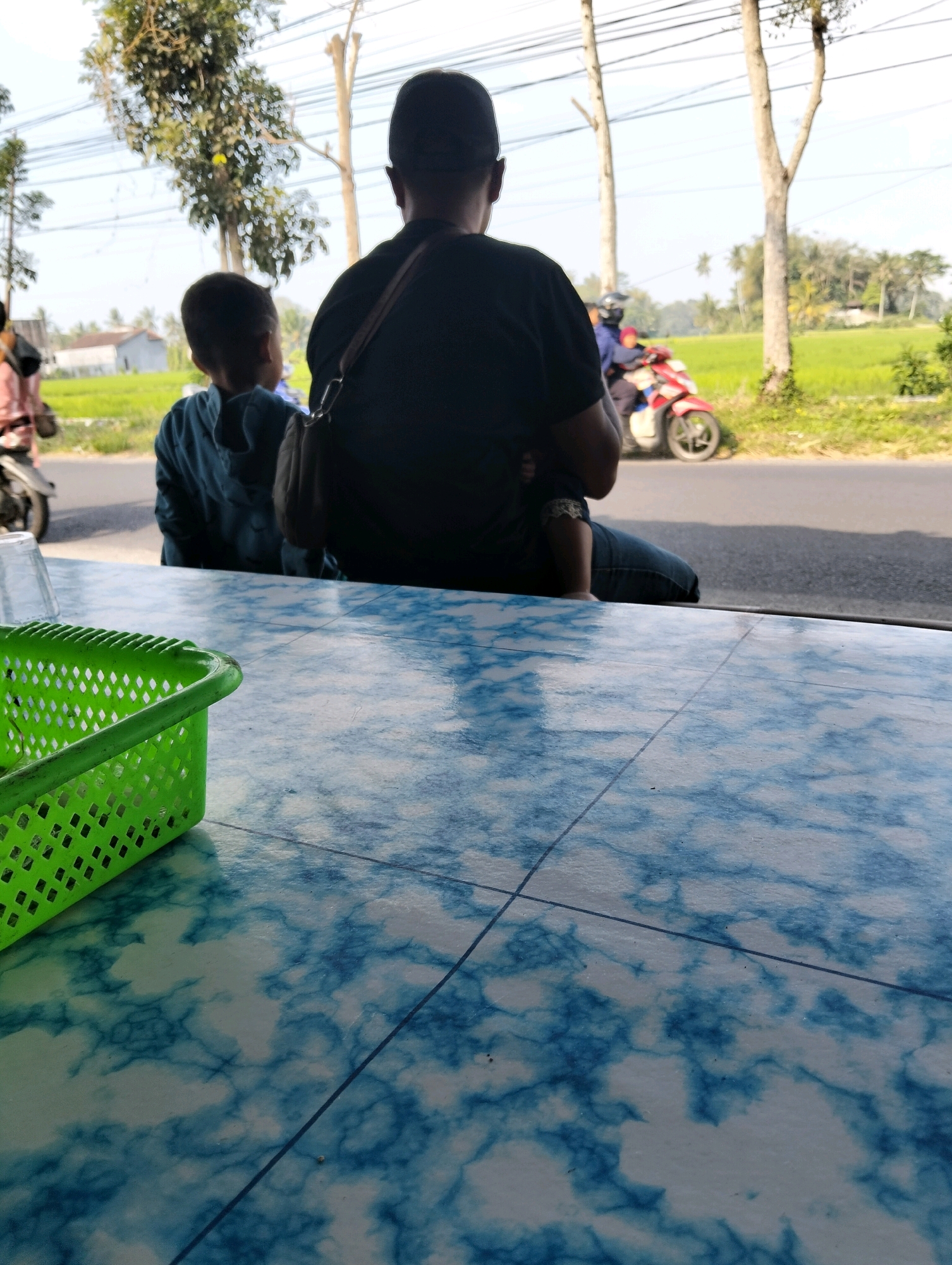 The image captures a scene from a roadside in Tegalarum, Indonesia. In the foreground, a blue and white marbled table is visible, with a green plastic basket on its left edge. The background shows two individuals, an adult and a child, from behind, seated on what appears to be a bench or a raised platform, facing away from the camera. The adult is wearing a dark t-shirt and jeans, with a bag slung over their shoulder. The child is wearing a blue hooded jacket. Beyond them, a paved road runs horizontally across the frame. A red and white motorcycle with two riders passes by on the road. To the right of the road, a vibrant green rice field stretches into the distance, bordered by trees and foliage. Power lines are suspended in the sky above. The overall impression is of a casual, outdoor setting, likely a small eatery or rest stop situated along a rural road in the specified Indonesian location.