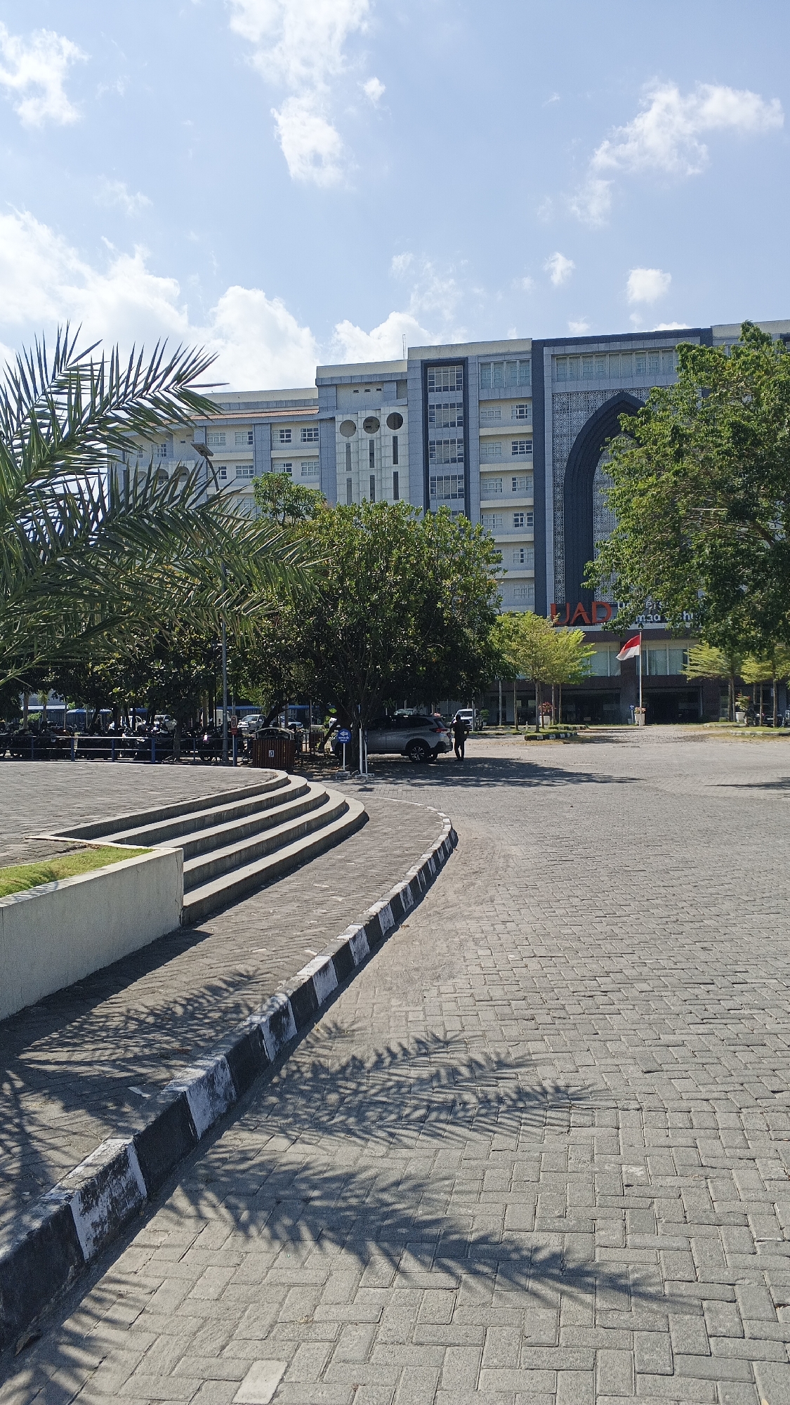 This is a full, medium-shot, eye-level, outdoor photograph taken on a sunny day. The image depicts a paved courtyard area in front of a large, modern building complex. The building has a light blue facade with rows of windows and decorative architectural elements, including a large arched entryway with intricate patterns. A red and white flag, likely the Indonesian flag, is visible on a flagpole in front of the building.
In the foreground, a curved paved area made of gray interlocking bricks dominates the lower half of the image. To the left, a series of concrete steps ascend a raised platform. A low, white concrete wall borders the grass on the platform. Palm fronds cast dappled shadows across the paved courtyard.
Several trees, including palm trees and other leafy trees, are scattered throughout the scene, providing shade. In the middle ground, a silver SUV is parked, with two individuals standing near it. A trash can is also visible. In the background, beyond the trees, are more of the building's structures and what appears to be a parking area with several motorcycles. The sky is blue with scattered white clouds.
Based on the architectural style of the building, which features Islamic geometric patterns and a prominent arched entrance, and the presence of the Indonesian flag, the location is likely a public institution or educational facility in Indonesia. The specific mention of Banguntapan, Indonesia, places this scene in the Bantul Regency of the Yogyakarta Special Region. The building's design suggests it could be a university or a government administrative center.