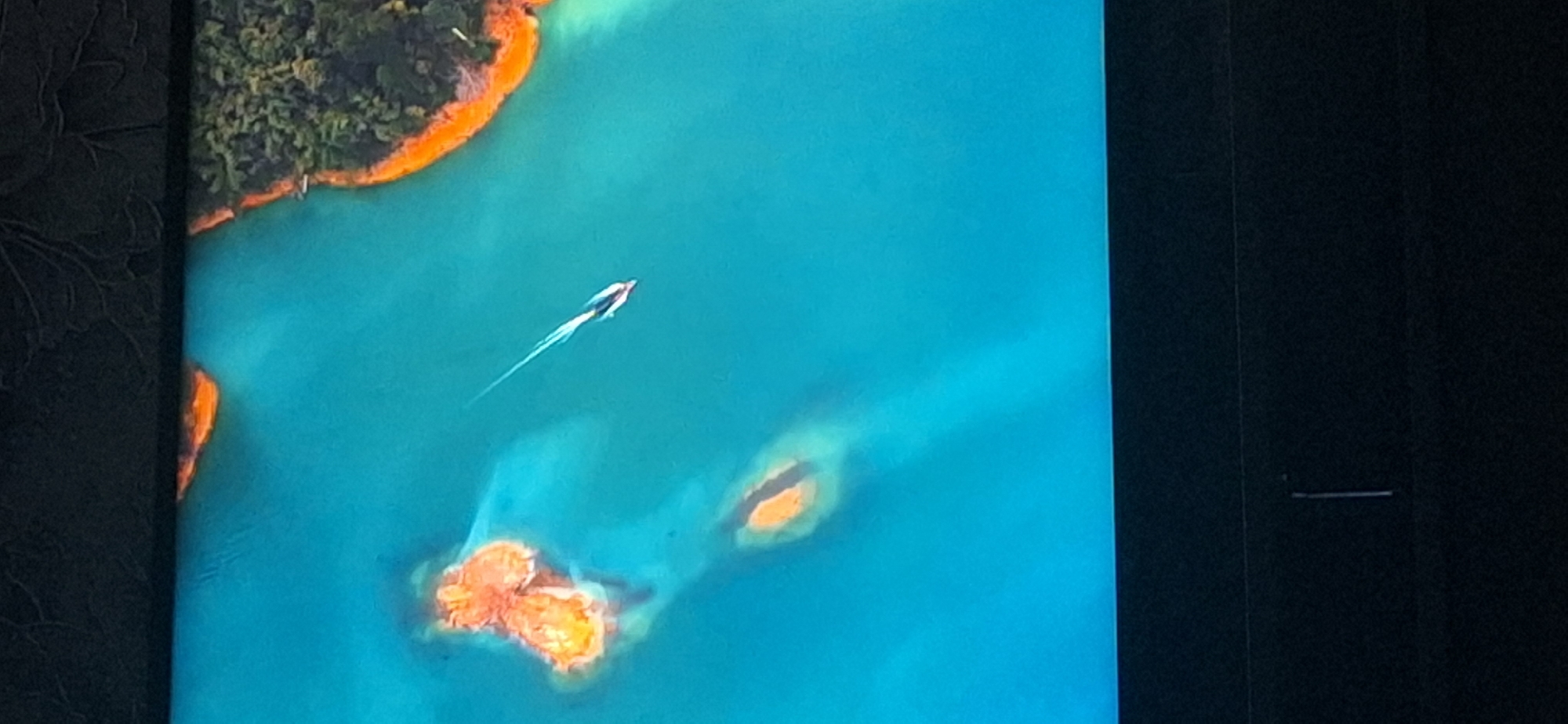 The image shows an aerial view of a body of water with a small boat moving through it. There are a few small islands with orange-colored edges and some vegetation on one side. The water is a vibrant turquoise color. The bottom portion of the image shows a dark area, possibly a screen or frame, with a thin vertical line in the center. The scene is not identifiable as being in Bila Tserkva, Ukraine, or any specific city.