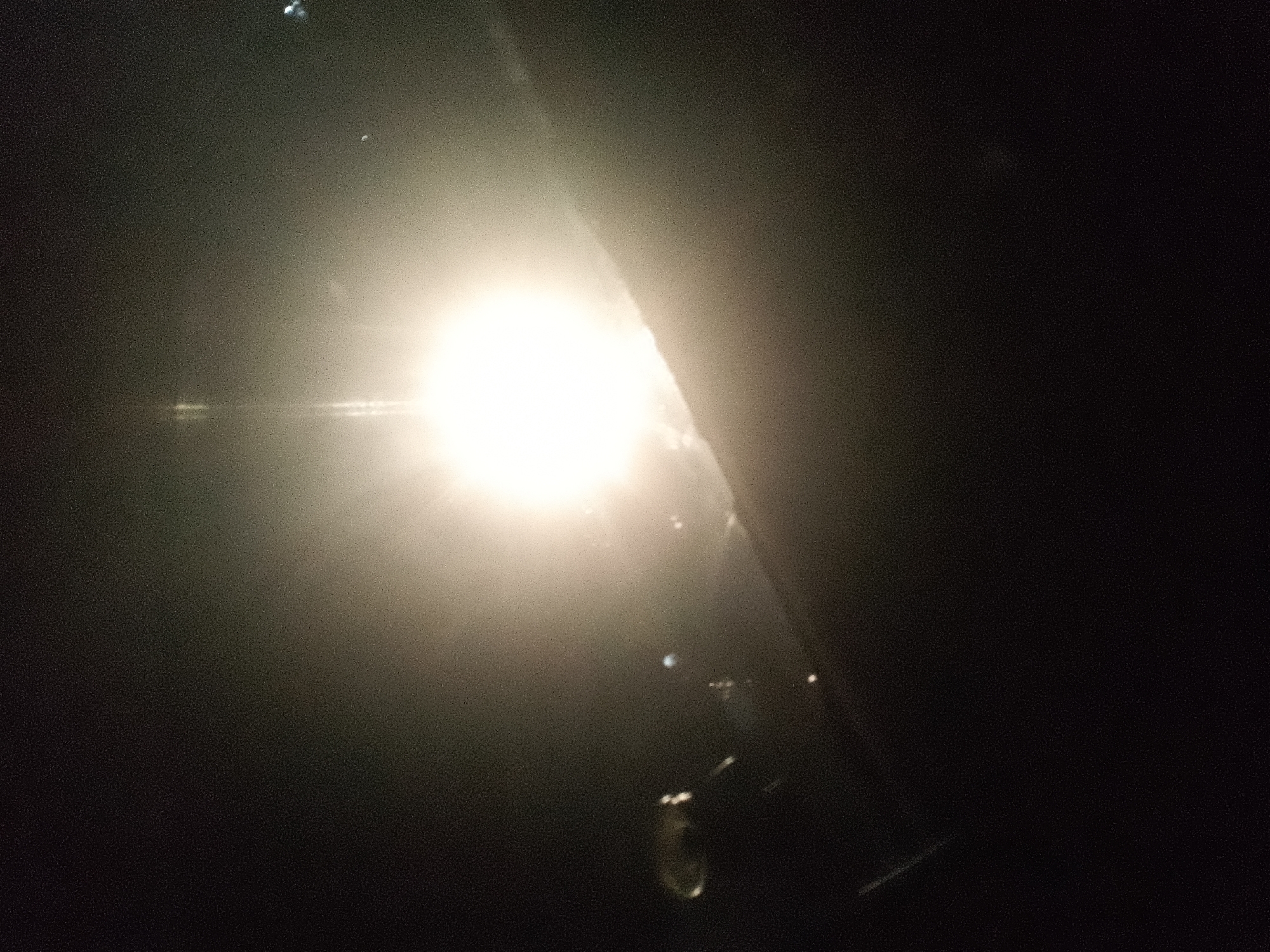 The image displays a bright, diffuse light source in a dark environment, suggestive of a strong light such as a spotlight or the sun obscured by fog or haze. The light emanates from the left side of the frame and extends across the center, creating a luminous halo. There are faint reflections or glints of light visible within the illuminated area and to its right. The background is predominantly dark, with some indistinct shapes and gradations in tone, hinting at a larger, unlit space or obscured objects. The overall impression is one of low visibility with a single dominant light source. The image was taken in Jalingo, Nigeria.