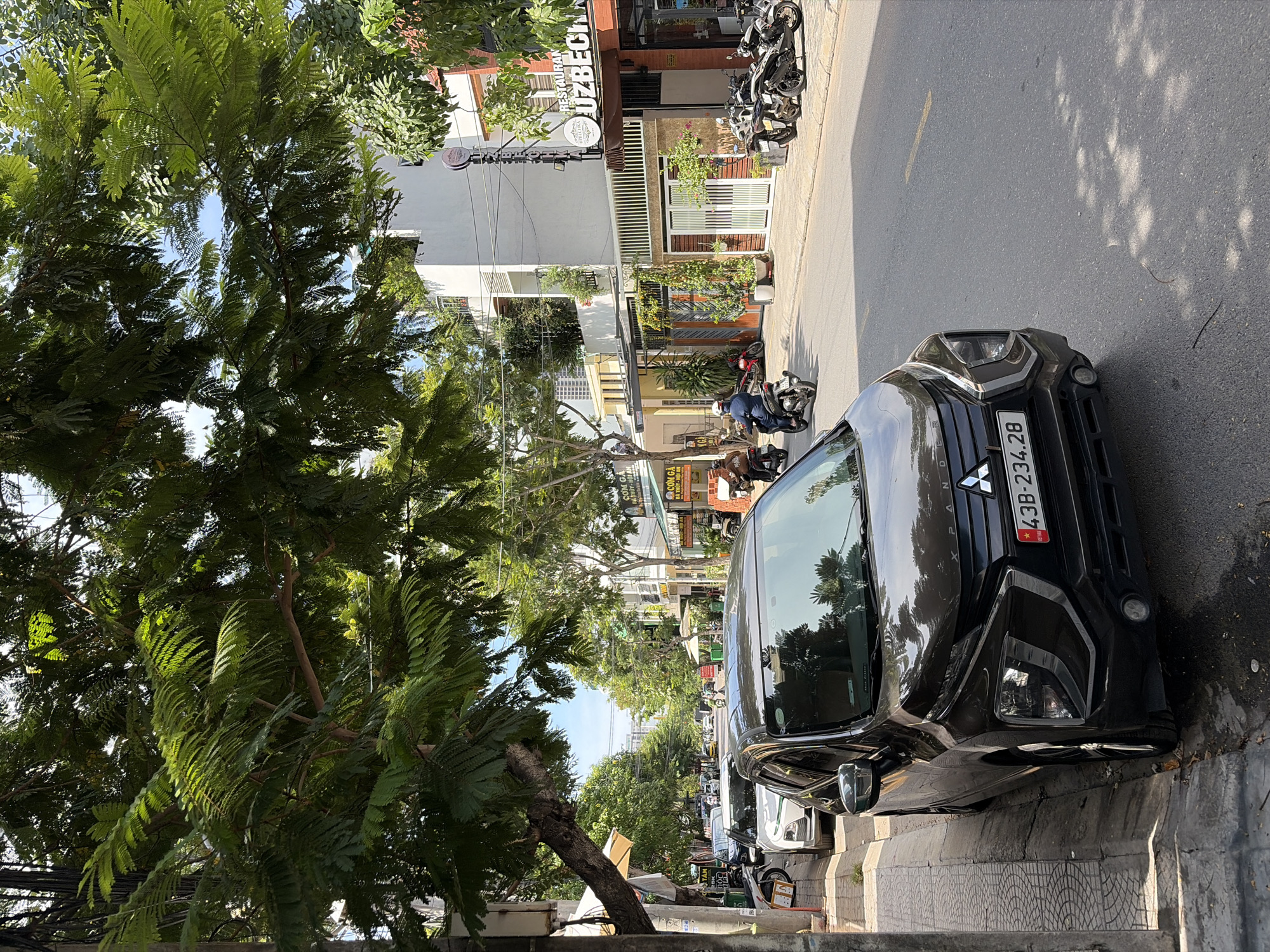 A street scene in Da Nang, Vietnam, shows a black Mitsubishi Xpander parked on the side of the road, its front end partially obscuring a sidewalk. The car has a license plate reading "43B-234.28". Across the street, several motorcycles are parked, and two individuals are riding them. Buildings line the street, featuring businesses with signage, including one that reads "RESTAURANT UZBECH". Lush green trees with dense foliage overhang the street, casting dappled shadows on the asphalt. The sky is visible through the leaves, appearing bright and clear. The overall environment suggests a sunny day in an urban setting.