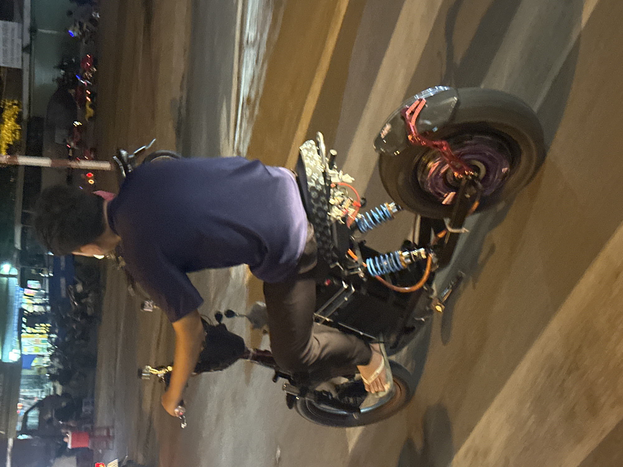 The image depicts a person riding an electric scooter at night on a street that appears to be in Bac Nha Trang Ward, Vietnam. The scooter is dark-colored with a battery pack visible underneath the seat, and it has a decorative pattern on the seat itself. The rider is wearing dark pants and a dark blue shirt, and is using white flip-flops on their feet. The scooter has bright blue and orange suspension components. The street is wet and reflective, suggesting recent rain or a humid environment. In the background, there are other vehicles and what appears to be a commercial area with illuminated signs, typical of a city street at night. The perspective is from behind and to the side of the rider, capturing the motion and the urban nighttime setting.