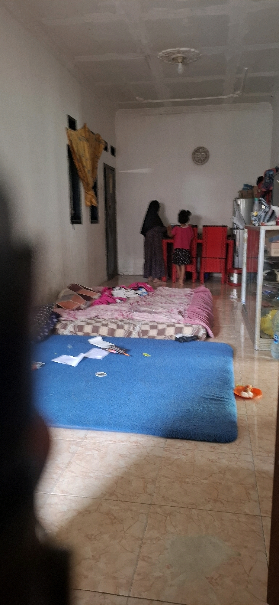 The image depicts an interior scene, likely a living area or bedroom, in a residence located in Ciharashas, Indonesia. Two individuals are present: an adult wearing a dark hijab and a long patterned dress, and a child in a pink shirt and patterned skirt. They are standing near a red table and chairs. In the foreground, there are two floor coverings: a pink and brown patterned mattress or bedroll, and a large blue mat. Papers and pens are scattered on the blue mat. A shelf unit with various items is visible on the right side of the image. The room has white walls and a drop ceiling with a light fixture. The flooring appears to be tiled. The overall impression is of a lived-in domestic space.