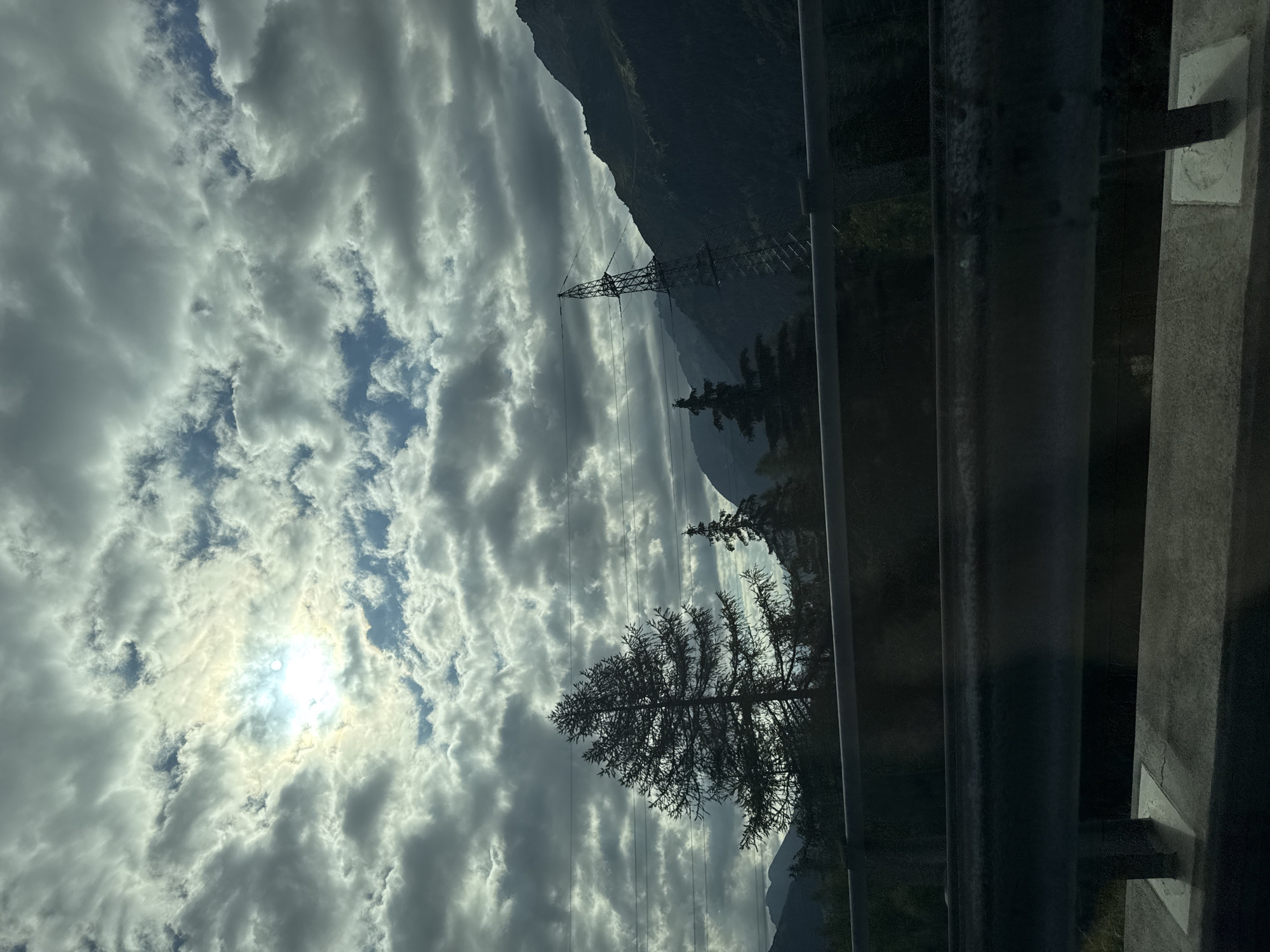 The image depicts a scenic view of the Swiss Alps near Airolo, Switzerland, under a dramatic sky filled with clouds. The sun is partially visible through a break in the clouds, casting a bright, diffused light. In the foreground, a metal guardrail runs horizontally across the frame, obscuring the lower portion of the landscape. Behind the guardrail, silhouetted pine trees stand tall against the backdrop of dark, forested mountains. A tall, metallic power pylon with wires extends upwards from the mountainside on the right, indicating human infrastructure within the natural setting. The overall impression is one of a vast, natural landscape characterized by mountainous terrain and an overcast sky.
