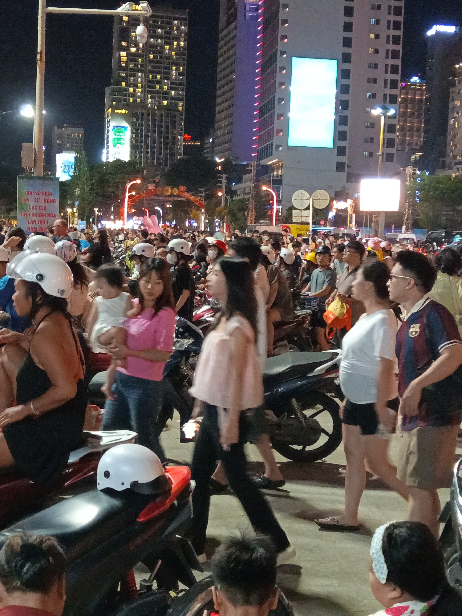 The image captures a bustling night scene in Nha Trang, Vietnam, likely on a street with significant pedestrian and vehicular traffic. Several tall, modern buildings dominate the background, some illuminated with colorful lights and large digital screens, suggesting a commercial or entertainment district. A street vendor's cart with a prominent "Maxim" sign is visible.
In the foreground and midground, a dense crowd of people are gathered, many on or around motorcycles. The people are dressed in casual, everyday attire, with varying ages represented. Some individuals are wearing helmets, either on their heads or placed on motorcycles. A child is being held by an adult, and other individuals appear to be walking, standing, or interacting within the crowd. The atmosphere is lively and appears to be a public gathering or event. The street surface is visible, showing a slightly textured pavement. The lighting is primarily artificial, from the buildings, streetlights, and vehicle headlights, creating a nighttime ambiance. The overall impression is one of a vibrant urban environment with a strong sense of community activity.