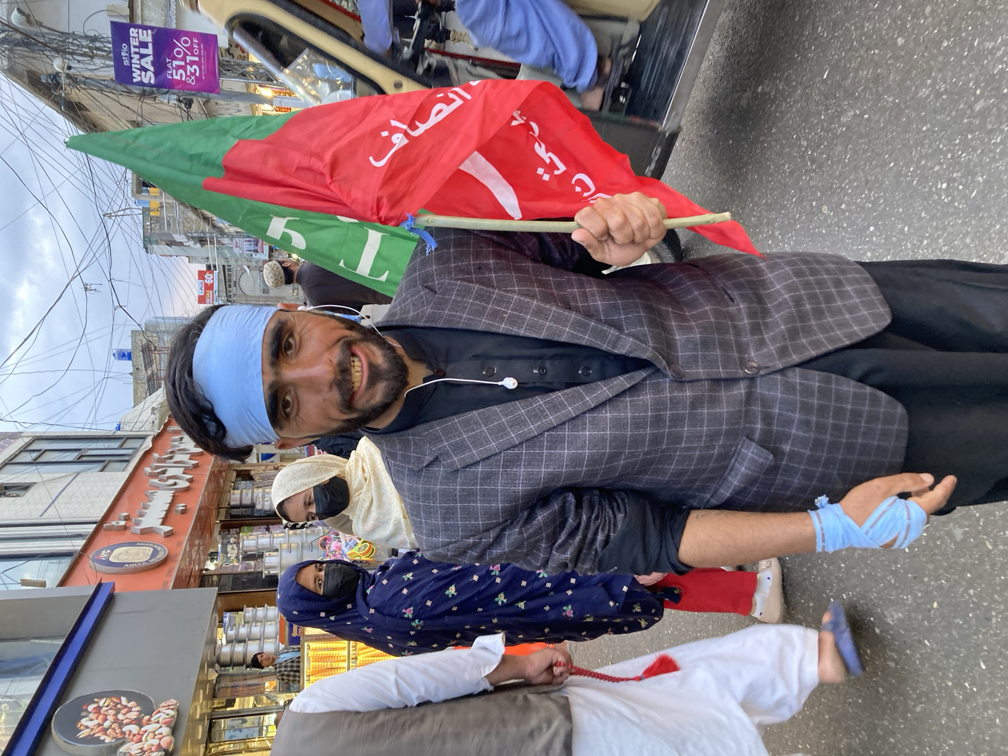 Here's a detailed description of the image:
**People:** A man is prominently featured, holding a green and red flag with Arabic text. He has a blue headband and appears to be wearing a checkered blazer over a black shirt. He has visible earphones. His right hand is bandaged. Several other individuals are visible in the background, including two women wearing masks and headscarves. A person in white is also visible further back.
**Subjects:** The main subject is the flag being held by the man. The other subjects include the building, shop signage, and other pedestrians.
**Objects:** Notable objects include the green and red flag, the man's blazer, a storefront with a sign "ABDUL", a "Winter Sale" banner.
**Actions:** The primary action is the man holding and displaying the flag. Other people are walking in the background.
**Interactions:** The man is engaging directly with the camera, seemingly presenting the flag. Other people appear to be walking about their business.
**Environment:** The scene appears to be a street setting in an urban area. There are buildings with storefronts, and power lines visible in the sky. Signage indicates a Winter Sale, suggesting a specific time of year. Based on the text on the flag and signs in the background, the location is likely Quetta City Tehsil, Pakistan.