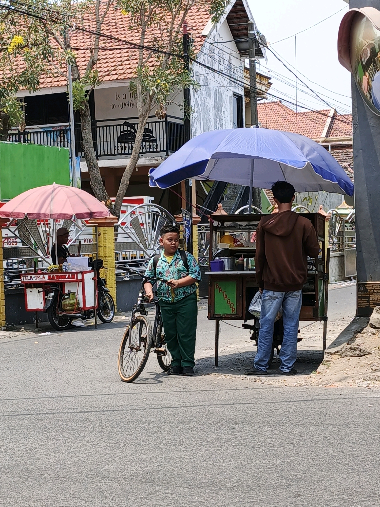 This is a street-level, eye-level, full-shot image captured in the city of Ketegan, Indonesia. On the right side of the frame, a young boy with short black hair is standing next to a bicycle with a light-colored frame and wheels, wearing a green patterned shirt and green pants. He is looking to the right. Behind him, a vendor is operating a food cart under a large blue umbrella. The cart has a dark metal frame and green panels with text. Another food cart is visible to the left, under a pink umbrella, with a vendor in a brown cap wearing a dark shirt. This cart is attached to a motorcycle. The street is paved, and a sidewalk with vegetation is visible in the background, along with buildings with terracotta tile roofs. The sky is bright and overcast.