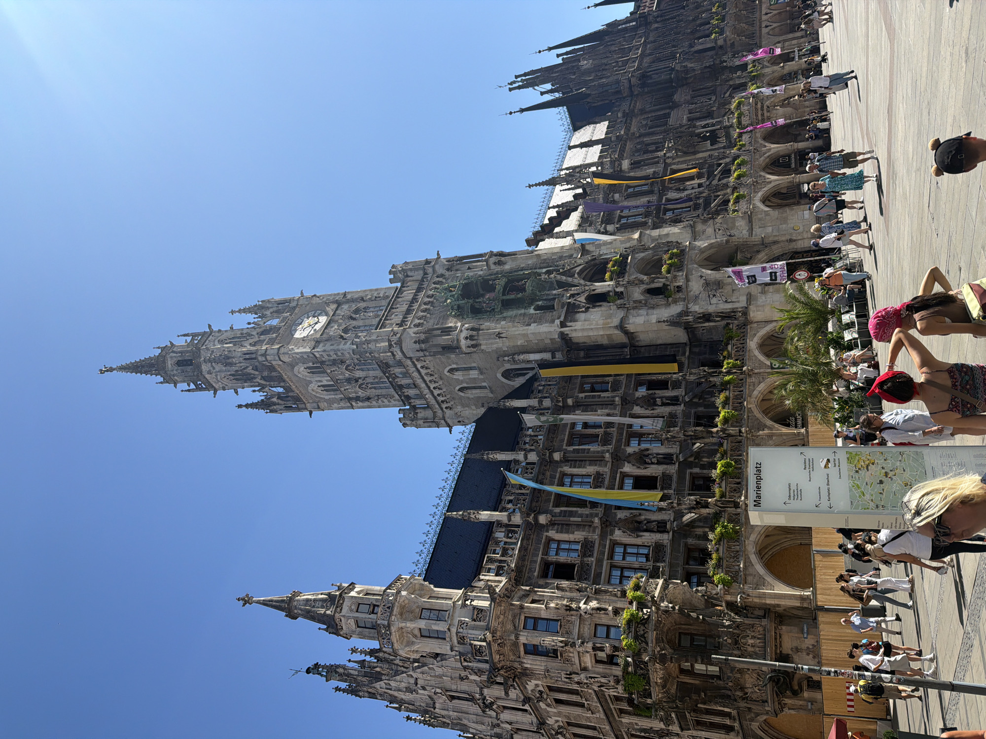 The image captures a sunny day at Marienplatz in Munich, Germany, a bustling public square known for its impressive architecture and vibrant atmosphere. Dominating the scene is the New Town Hall (Neues Rathaus), a magnificent Gothic Revival building characterized by its ornate facade, intricate spires, and a prominent clock tower. The building's elaborate stonework, detailed statues, and arched walkways are clearly visible, showcasing its historical grandeur.
In front of the New Town Hall, a diverse group of people are seen strolling through the square. Tourists are present, some with backpacks, suggesting they are exploring the city. Families and individuals are casually walking, conversing, and enjoying the pleasant weather. Some individuals are looking at a signpost that indicates directions to various landmarks, including "Marienplatz," "Rathaus," and "Deutsches Museum," further emphasizing the location as a central hub. Several flags, including the Ukrainian flag, are displayed on the building, adding a splash of color to the scene. Palm trees and greenery are interspersed throughout the square, providing a touch of nature amidst the urban landscape. The sky is a clear, bright blue, indicating good weather conditions. The overall impression is one of a lively and historically significant urban space.