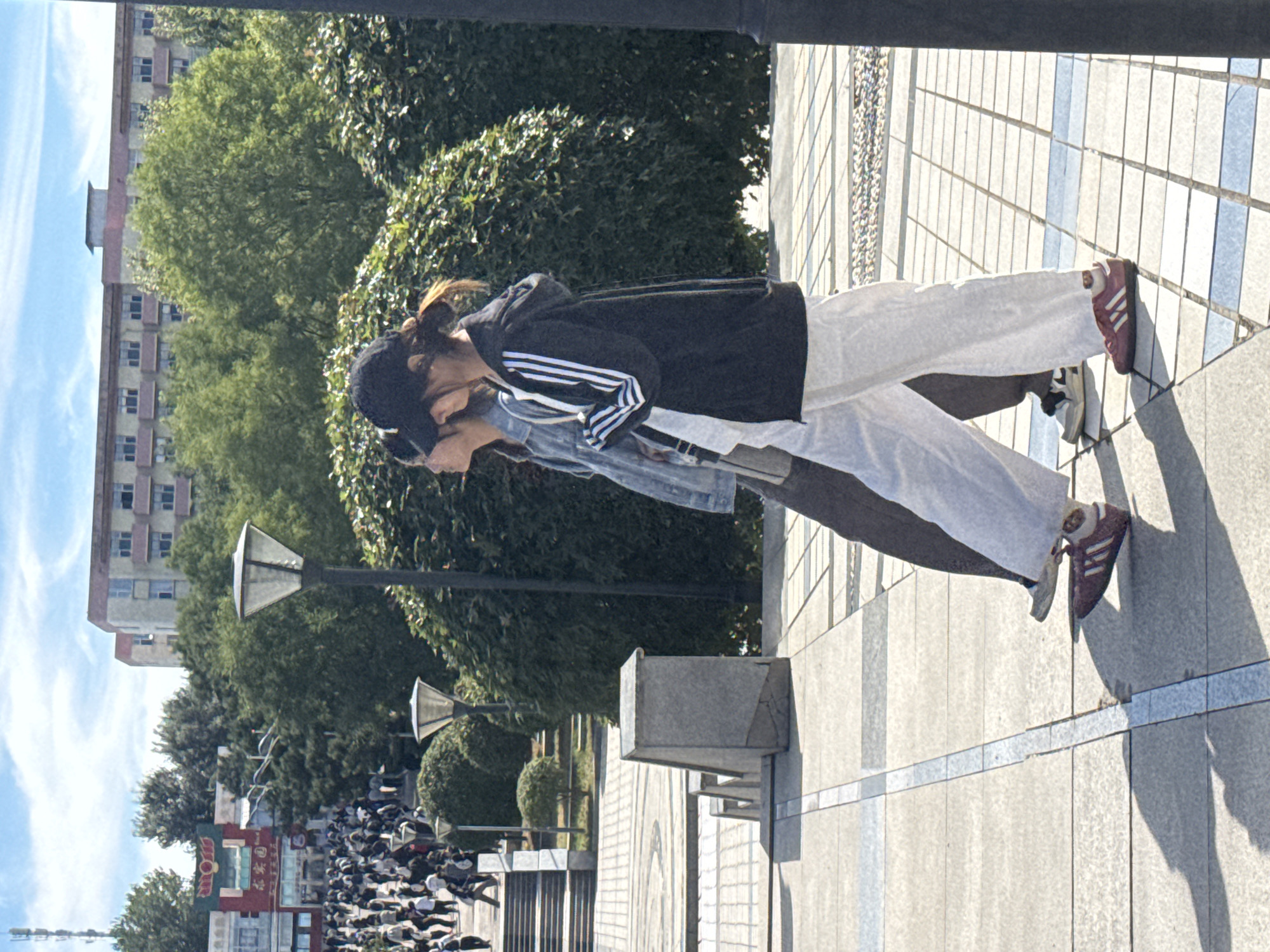 This image captures a sunny day scene on a paved walkway in Guta, China. In the foreground, two young women are walking side-by-side. The woman on the right is wearing a black zip-up hoodie with white stripes on the sleeves, white wide-leg pants, and maroon sneakers with white stripes. She has her hair pulled back in a ponytail and is wearing a black baseball cap. The woman on the left is wearing a denim jacket over a light-colored shirt and dark pants. Her hair is also pulled back. They are walking on a wide, light-colored tiled path with darker grey border lines. Their shadows are cast on the ground to their left.
In the mid-ground, a stone structure and a lamppost are visible. Beyond them, a densely vegetated area with large, rounded bushes is present. Further back, a crowd of people are walking up a wide set of stairs. To the left of the stairs, there are buildings with colorful facades and signs, suggesting a commercial area. In the background, several multi-story buildings with light-colored exteriors and numerous windows can be seen, as well as lush green trees. The sky above is blue with scattered white clouds. The overall environment suggests an outdoor public space, possibly part of a university campus or a recreational area in Guta.