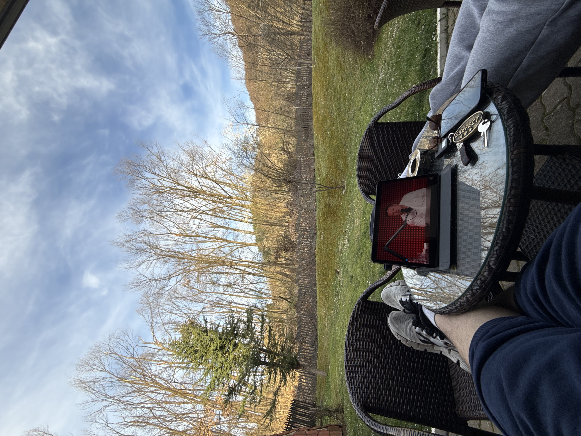 The scene is an outdoor patio setting, likely a backyard or garden. The camera is positioned at an angle, looking down at a person seated at a round glass-topped table. The person is wearing a pair of grey and black sneakers, dark blue pants, and grey sweatpants (partially visible). A tablet rests on the table, displaying an image of a man on a red background, possibly a video or a presentation. Also on the table are a pair of sunglasses, keys, and a smartphone. The patio is furnished with dark wicker chairs.
The background features a lawn, a wooden fence, and a row of trees with bare branches, typical of late autumn or early spring. The sky is partly cloudy with patches of blue visible.
No specific information indicates that the location is in the city of Etropole, Bulgaria. This detail requires additional contextual information or identifying features not present in the image.