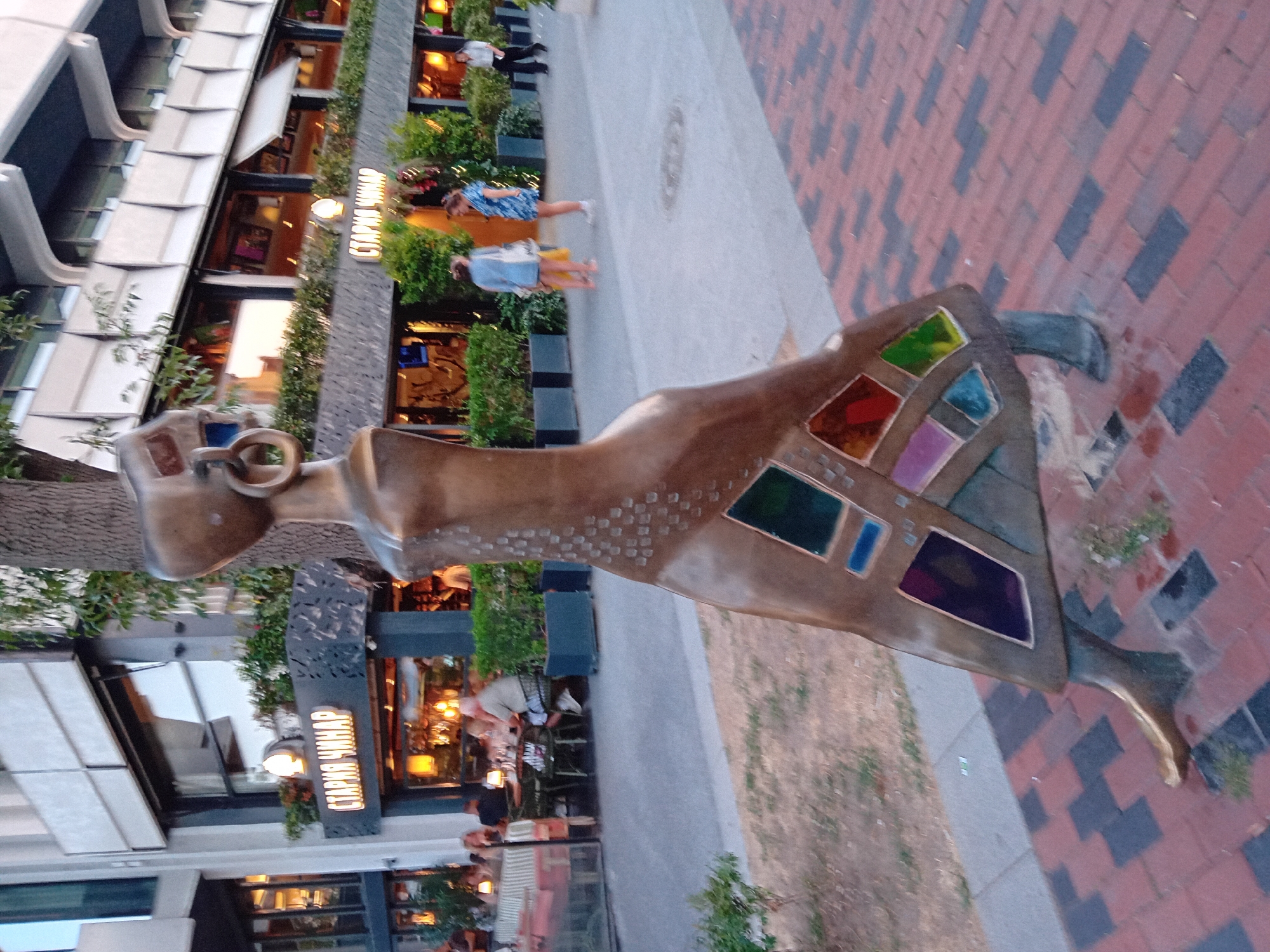 The image captures a statue of a woman, rendered in a bronze-like material with colorful glass inlays on its skirt, positioned on a brick-patterned walkway in the city of Varna, Bulgaria. The statue is located in front of a restaurant with outdoor seating. The restaurant's signage, written in Cyrillic, reads "СТАРИЯ ЧИНАР". In the background, people are seated at tables in the restaurant and walking along the pavement. The environment appears to be an urban setting with a building structure behind the restaurant.