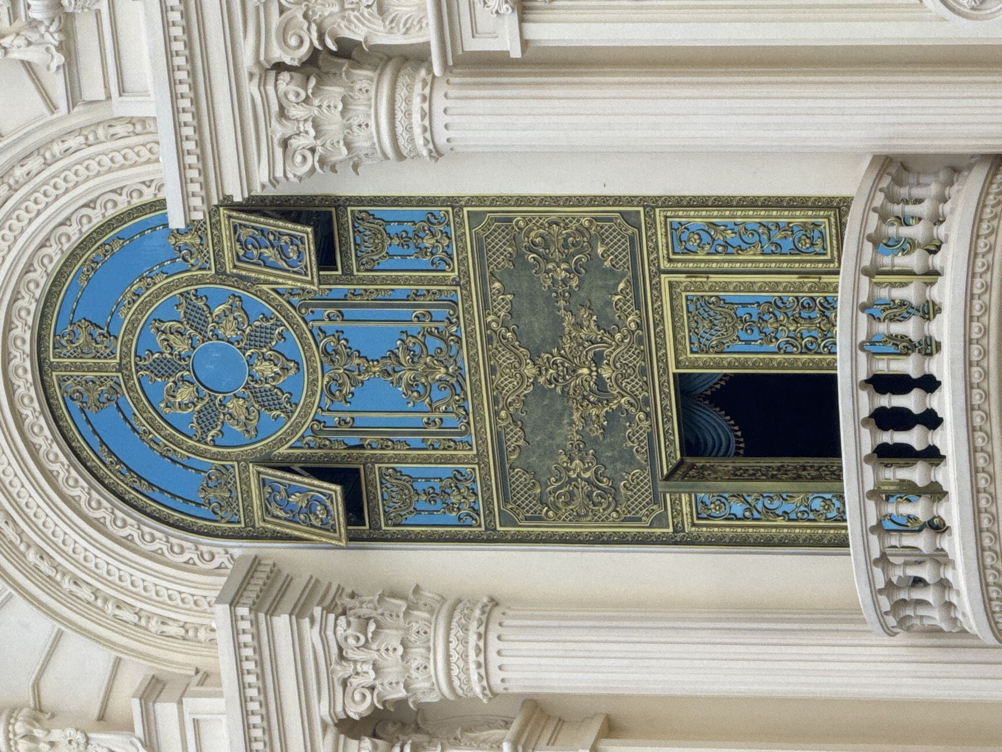 The image shows an ornate, antique window with intricate gold detailing against a vibrant blue background, situated on a cream-colored building. The window is framed by decorative architectural elements, including columns and a balustrade. The style of the building and the window suggests a European influence, possibly French colonial, which is common in Da Lat, Vietnam, due to its history as a French hill station. The building's architecture, with its classical columns and decorative moldings, indicates it's a significant structure, likely a historical landmark or a well-preserved colonial-era building in Da Lat. No people are visible in the image. The window is part of the facade of a large building, and the surrounding elements such as the columns and the balcony suggest it is on an upper floor. The lighting suggests it is daytime, with soft light illuminating the scene.