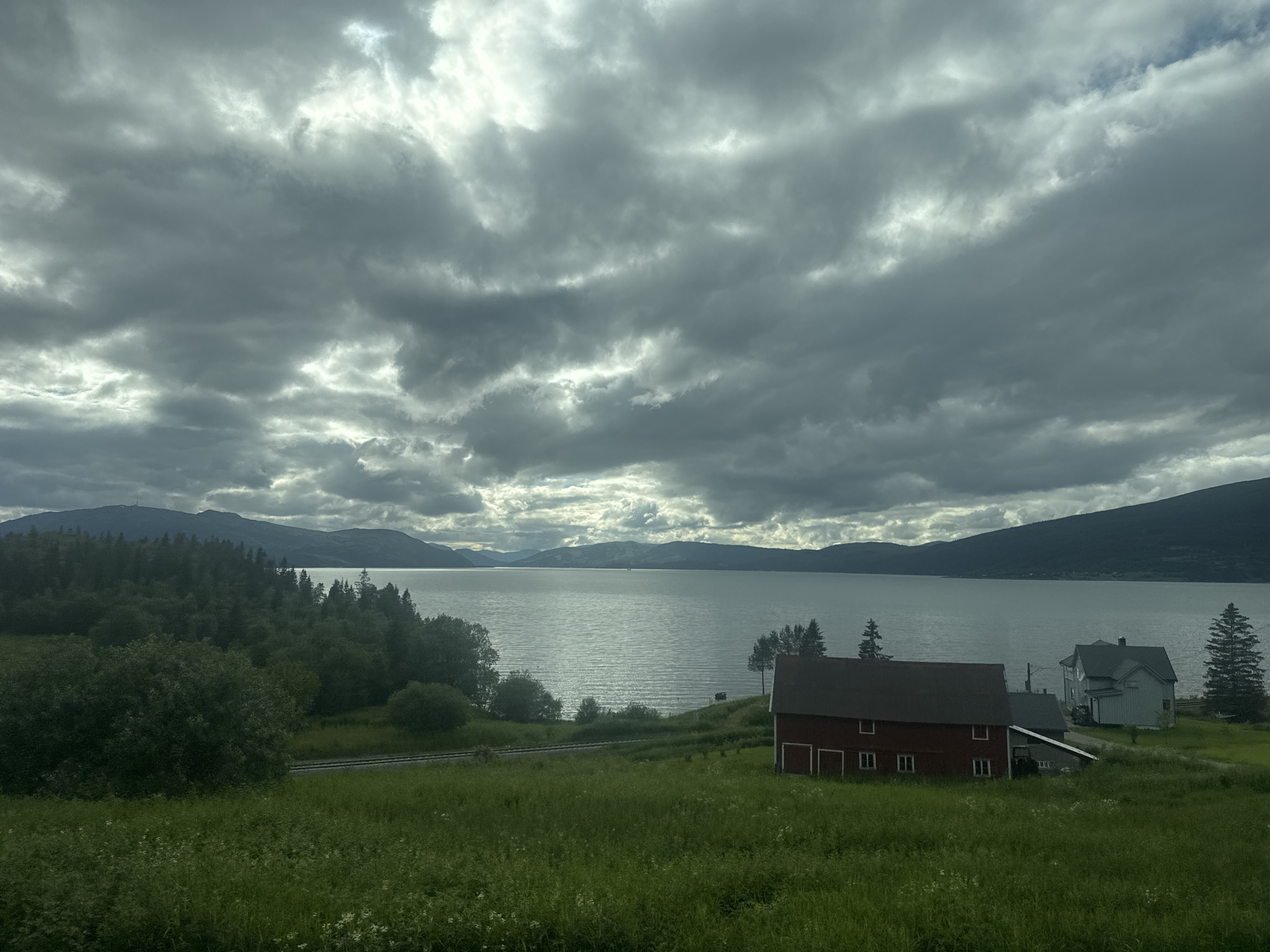 This landscape photo depicts a serene rural scene in Dalsgrenda, Norway, under a dramatic, cloudy sky. The foreground is dominated by lush green grass, with scattered white wildflowers. A dark red barn with white trim and multiple windows stands prominently on the right side of the frame. Adjacent to it, a lighter colored, multi-story house with a gray roof is visible, nestled amidst the greenery.
Beyond the buildings, a dense forest of evergreen trees lines the edge of a large, calm body of water. The water reflects the overcast sky, creating a silvery sheen. In the distance, rolling hills and mountains form the backdrop, their dark silhouettes softened by the atmospheric conditions. A faint glimmer of light breaks through the clouds over the water, suggesting it might be midday, despite the heavy cloud cover. The overall mood is one of peacefulness and natural beauty. No people are visible in the image. There is no visible text.