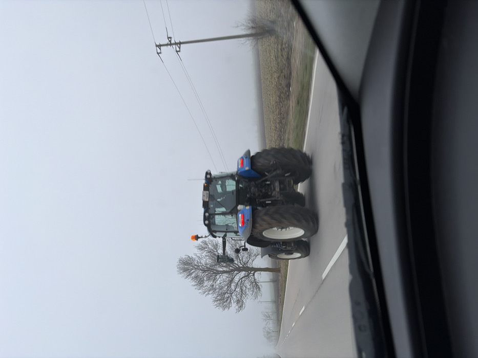 The image, captured from within a vehicle looking forward through the windshield, depicts a rural road in Ezerets, Bulgaria, under overcast and foggy conditions. A blue New Holland tractor is driving away from the camera in the right lane of a two-lane asphalt road. The tractor features large, dark-tread tires with white rims, and an amber warning beacon is mounted on its cab roof, which also displays a rear license plate. Two indistinct figures are visible within the tractor's cabin. The road surface is dark asphalt with a solid white line on the right shoulder and a dashed white center line. To the left, a line of bare deciduous trees runs parallel to the road, fading into the dense fog. To the right of the road, a dark, cultivated field is visible, extending towards a distant utility pole with power lines. The low visibility indicates significant atmospheric fog or heavy cloud cover.