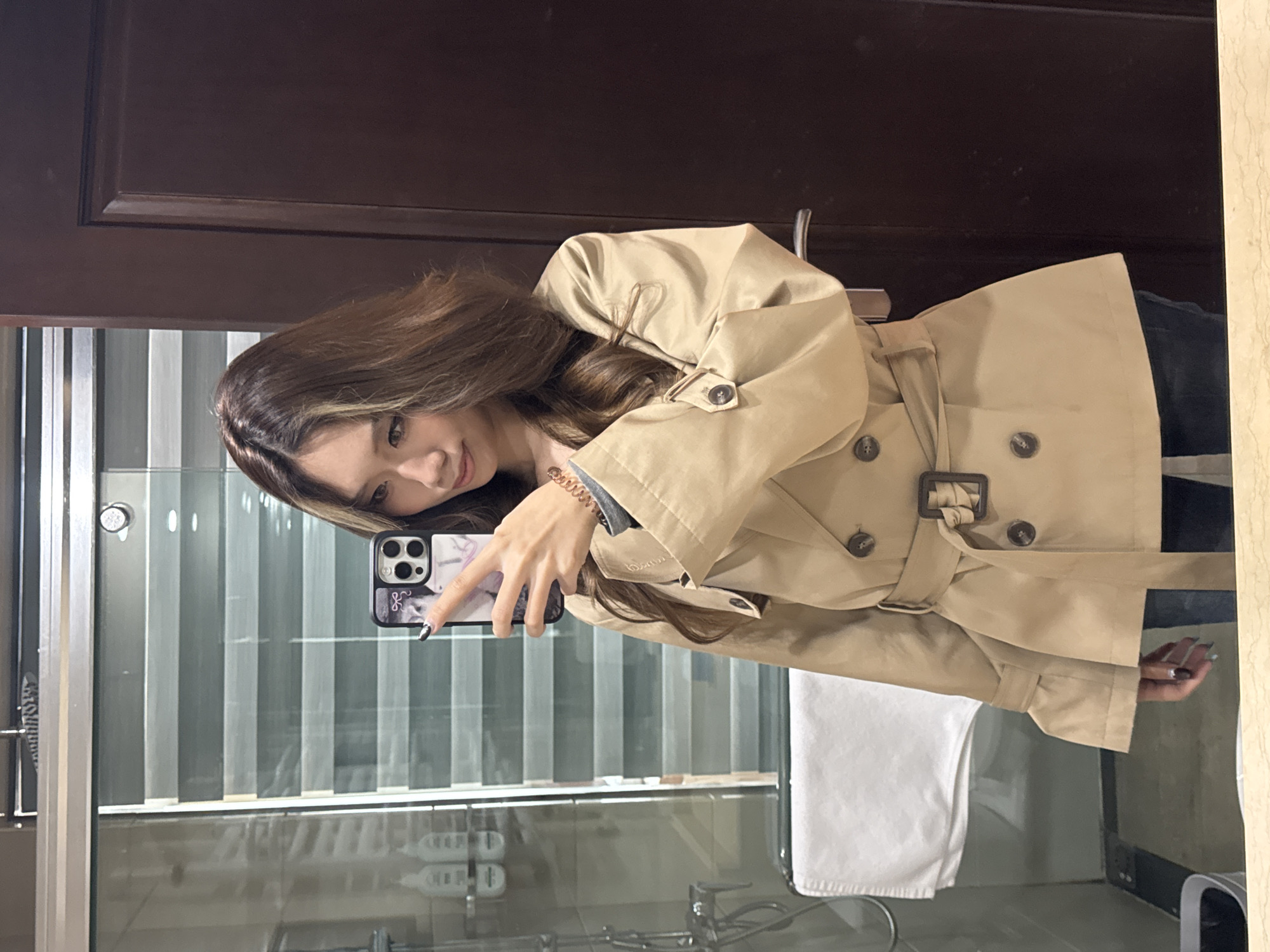 A young woman with light brown hair poses for a selfie in a bathroom. She wears a tan trench coat. The bathroom has a shower with glass doors and a white towel. The location is likely Lanzhou, China.