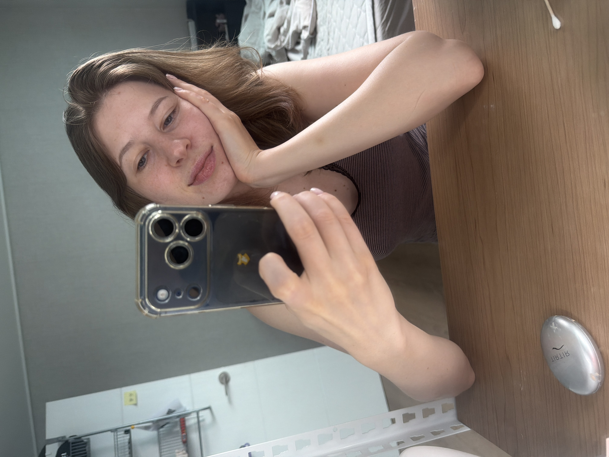 A young woman with light brown hair is taking a selfie in a room. She is leaning her head on her right hand, and her left hand is holding a smartphone. She is wearing a striped tank top. On the wooden table in front of her, there is a silver oval-shaped object and a cotton swab. In the background, a metal shelf unit is visible, as well as a white tiled wall and a door handle, suggesting a bathroom or utility area. A bed with grey linens is also present. The scene appears to be indoors, possibly in a residential setting in Dongducheon-si, South Korea.