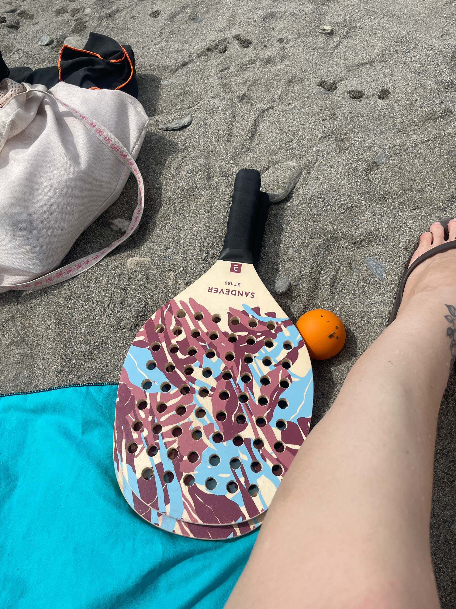 The image shows a beach scene in La Herradura, Spain. A paddleball racket and an orange ball are resting on the sand. A teal beach towel is visible to the left, and a light-colored canvas bag with a pink ribbon is in the background. A person's bare leg and foot wearing a flip-flop are also visible in the foreground. The sand is a mixture of fine grains and small pebbles, typical of the beaches in the Costa Tropical region. The sunlight suggests a bright, clear day, ideal for beach activities.