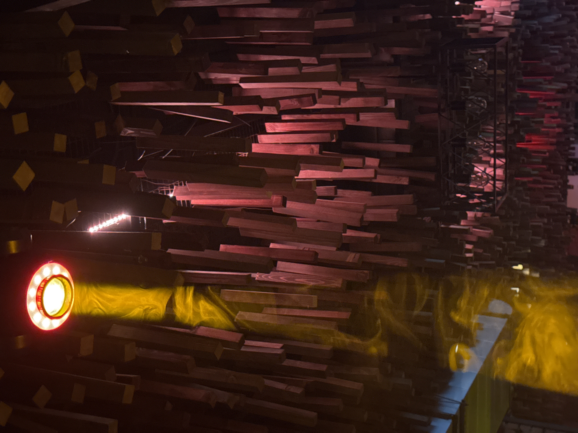 This image depicts an interior scene, reportedly located in Cheboksary, Russia, characterized by a unique, elaborate ceiling structure and dramatic lighting.
The dominant feature is the ceiling, densely covered with numerous irregularly hung, rectangular wooden blocks of varying lengths and orientations. These blocks are primarily a dark reddish-brown or maroon color. Some of the blocks, particularly those higher up and towards the left, have distinct small, light yellow square caps or ends, possibly indicating their mounting points or a decorative feature. The background behind and above the blocks is dark.
In the upper left quadrant, a prominent circular light fixture is visible. It features a bright red outer rim with several internal red lights and a central, intensely bright yellow light source. This fixture emits a strong, focused beam of yellow light that projects downwards and slightly to the left, illuminating the hanging wooden blocks and appearing to pass through a visible haze or smoke, creating undulating patterns in the light beam itself.
Towards the upper right, several smaller, reddish-pink light sources, possibly linear LED strips, are partially visible in the distance. In the lower-right portion of the ceiling, a black, open-cage style light fixture containing multiple clear, spherical bulbs is suspended. This fixture casts a reddish glow onto the blocks directly below it.
In the bottom left corner, a reflective surface, possibly a window or screen, exhibits a distinct blue hue and reflects some of the yellow light beam and ambient light.
No people are present in the scene. The overall environment suggests a modern, atmospheric venue such as a restaurant, bar, or club, designed with intricate decorative elements and dynamic lighting.