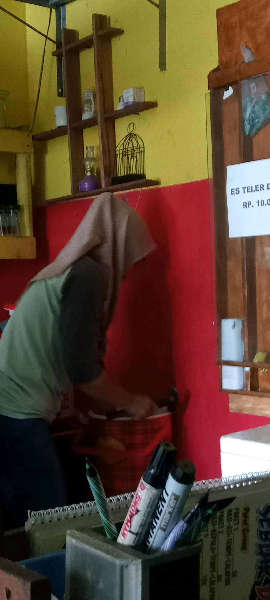 A person wearing a light brown hijab and a green shirt is in a room with yellow and red walls. They are leaning over a red checkered basket, appearing to be placing or retrieving something from it. In the background, a wooden shelf with various items, including a small birdcage, is mounted on the yellow wall. A sign on the right side of the image reads "ES TELUR" and "RP. 10.00". In the foreground, a desk organizer holds pens and markers, including one with "SNOWMAN" written on it. The room appears to be an indoor establishment, possibly a food stall or shop, given the menu sign. The context of the sign and the overall setting suggest the location is within Pondok, Indonesia.