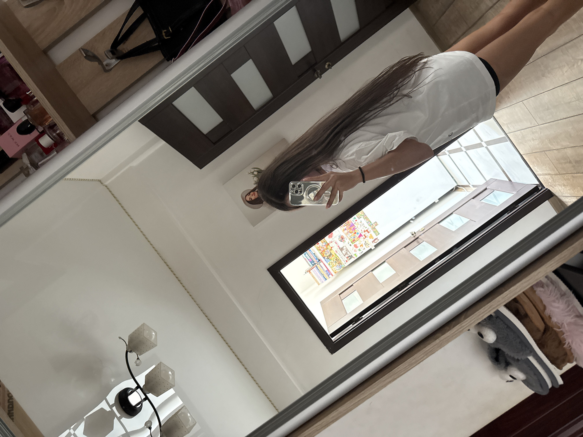 The image shows a young woman in a white t-shirt and black shorts taking a mirror selfie. She has long, dark brown hair and is holding a smartphone. In the background, a closet with shelves of perfume bottles and a black handbag are visible. To the left, a ceiling light fixture and slippers can be seen on a shelf. The scene appears to be indoors, likely a bedroom or dressing area, with tiled flooring and wooden doors. No specific location in Chișinău, Moldova can be determined from the image.