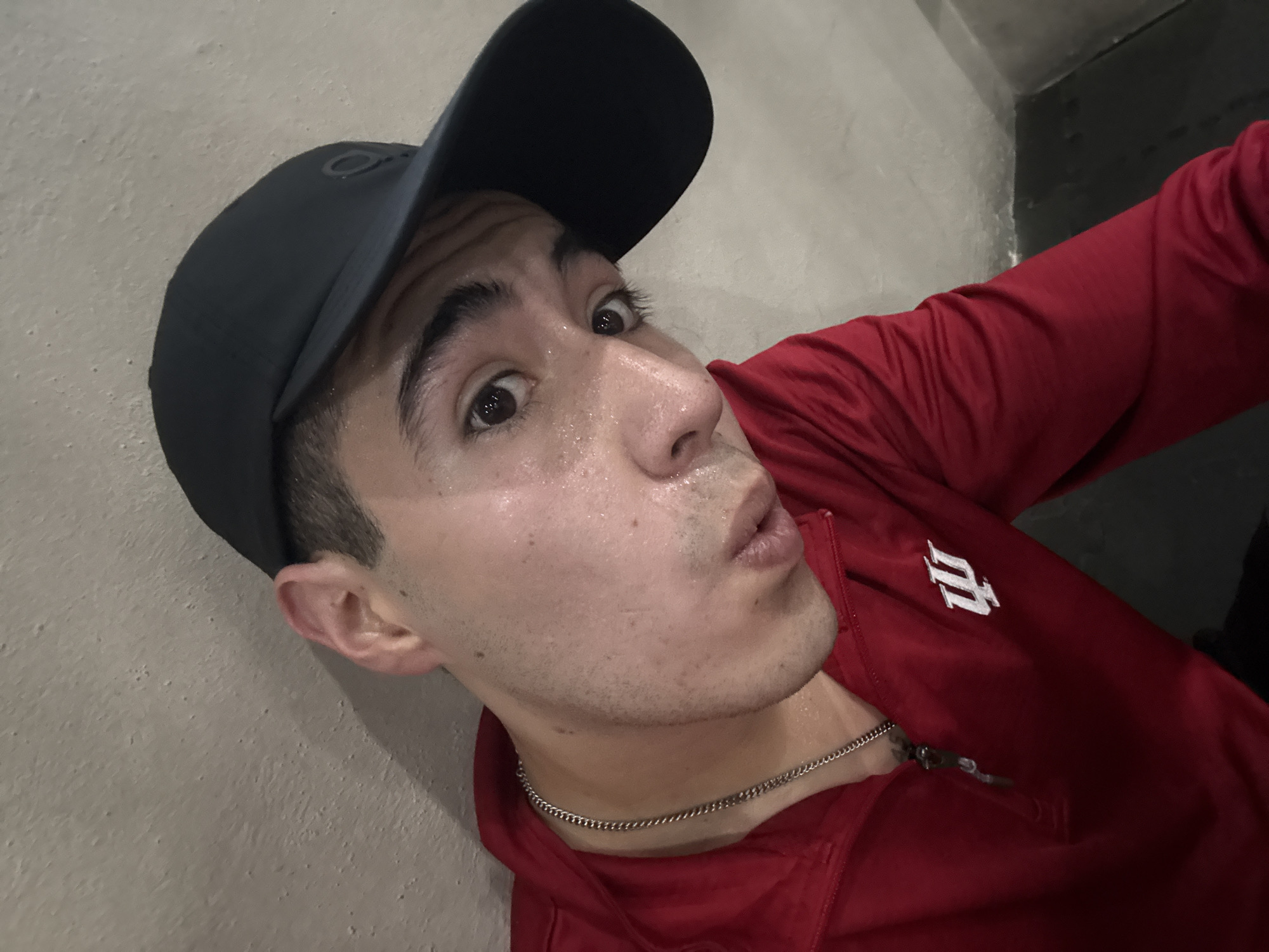 A selfie taken at an indeterminate location. The subject is a young adult male wearing a black baseball cap and a red jacket with a white "I" logo, suggesting affiliation with Indiana University. He is wearing a silver chain. His skin is damp, likely from perspiration. The background consists of a rough, gray wall and a dark corner. The location is unknown. There is no evidence to suggest the image was taken in San Bernardino Tlaxcalancingo, Mexico.