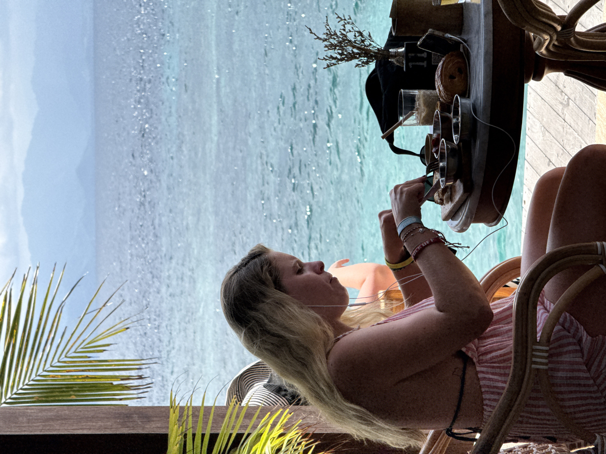The image shows a blonde woman sitting on a wicker chair at a waterfront table in what appears to be Nusa Penida, Indonesia, suggested by the tropical vegetation and ocean setting. She is wearing a striped red and white top, has multiple bracelets on her left wrist, and is wearing earphones. She is looking down at a breakfast tray on the table in front of her and appears to be eating with a fork in her right hand and a knife in her left. The tray contains a variety of breakfast items including pastries, fruit, and eggs. Behind the table and the woman is a view of the ocean. A palm frond partially obscures the view on the left side of the frame.