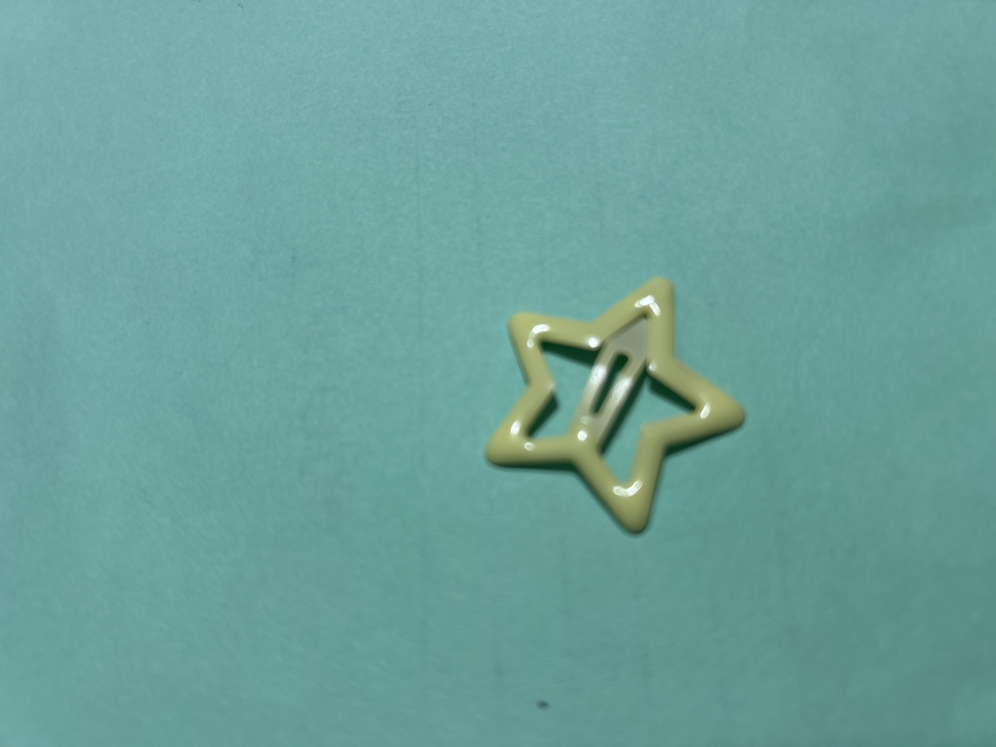 A pale yellow star-shaped hair clip is positioned in the center of the image against a teal background. The clip is made of plastic and has a hollow interior. The background appears to be a flat surface with a slight texture. There are no people or any other objects visible in the image. The location in the city of Дедемель, Kyrgyzstan is not determinable from the provided image.