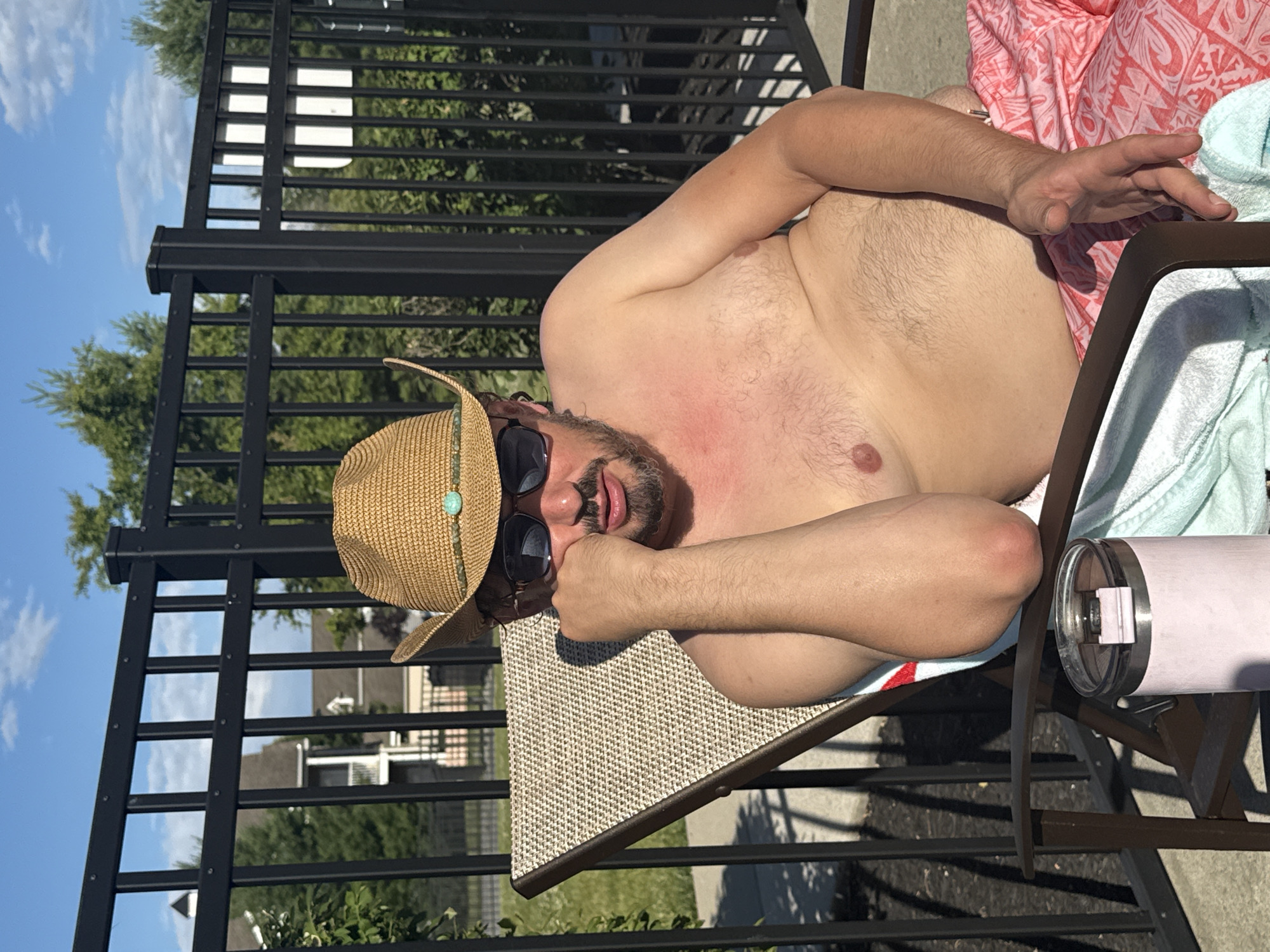 A Caucasian man wearing a straw cowboy hat and sunglasses reclines on a lounge chair by a fence and trees. He is shirtless and wearing patterned shorts. A pink tumbler is visible in the foreground next to a towel. The background shows houses and a blue sky with clouds. The man is resting his head on his hand.