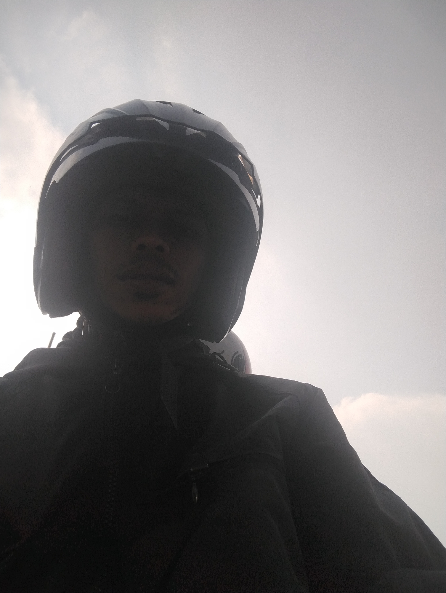 A low-angle, dimly lit, selfie-style photograph captures a person wearing a dark motorcycle helmet. The helmet covers most of the person's head and partially obscures their face, revealing only a mustache and a hint of the nose and mouth. The person is also wearing a dark jacket. The background is a bright, overcast sky with some wispy clouds. The image appears to be taken outdoors, possibly while riding a motorcycle. The lighting suggests it is daytime. The individual's face is largely in shadow due to the helmet and the angle of the sun.