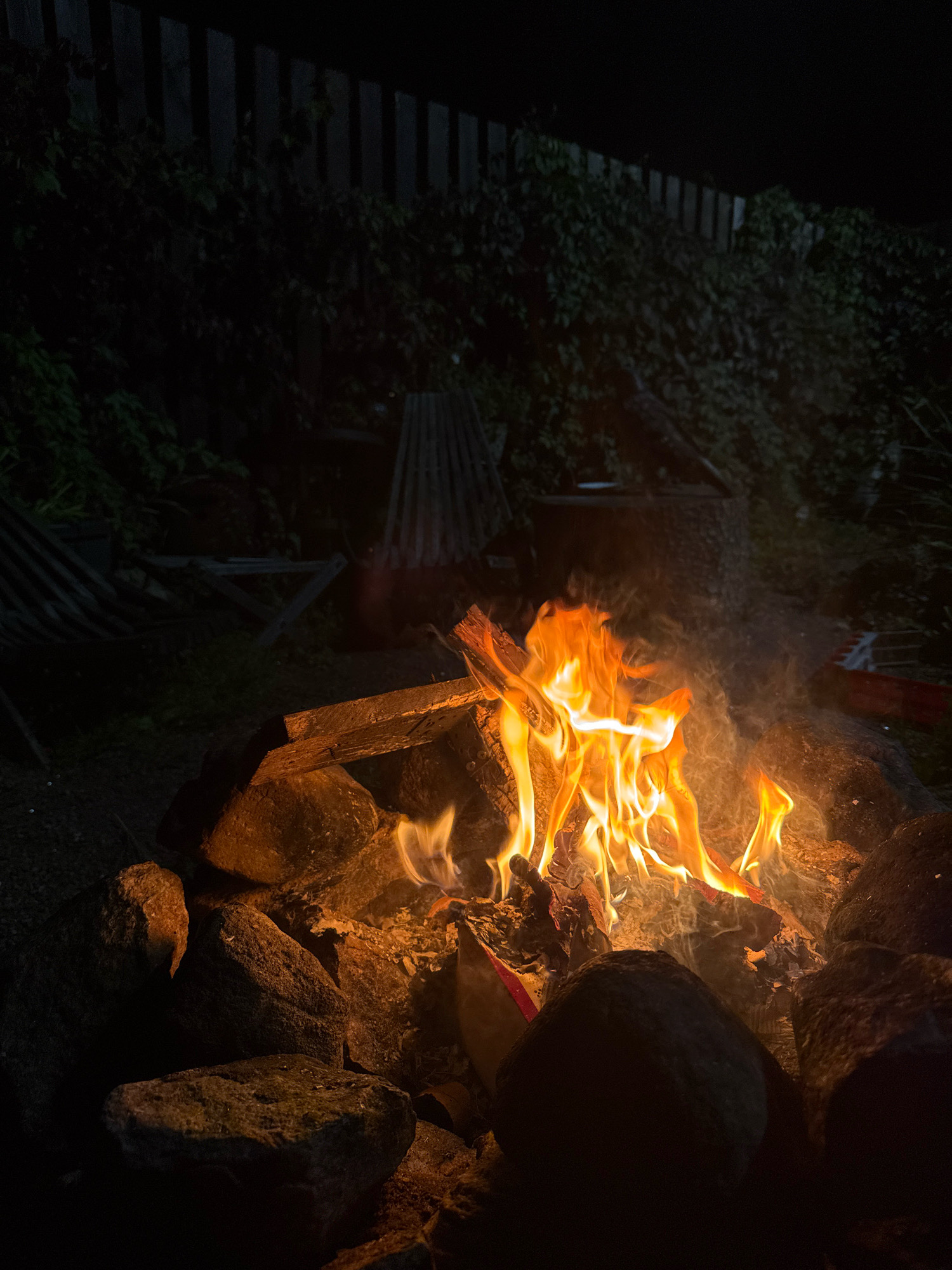 A campfire burns brightly in a backyard at night. The fire is contained within a ring of large, rough stones. Flames are actively consuming logs and emitting smoke. The background is dark, with a wooden fence visible in the distance, partially obscured by dense green foliage. In the mid-ground, there are some discarded pieces of wood or construction materials, and a faint suggestion of outdoor furniture. The overall setting appears to be a residential outdoor space, potentially in a suburban or rural area. The specific location in Kempelevo, Russia, is not visually evident from the image. There are no people present in the scene.