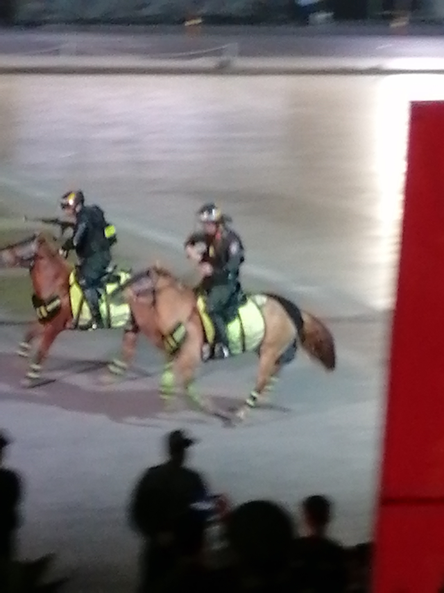 Two mounted police officers on horseback are visible in the image, which appears to be taken during a daylight event or parade. The officers are wearing helmets and dark uniforms with reflective yellow vests. The horses are adorned with yellow leg wraps and saddle blankets. The ground is paved, and a blurred red structure is present on the right side of the frame. In the foreground, several silhouetted figures are present, also appearing to be spectators. The background is out of focus, showing a greyish, outdoor environment. The scene depicts a law enforcement presence at a public gathering. There is no indication of the image being specifically located in Da Lat, Vietnam.