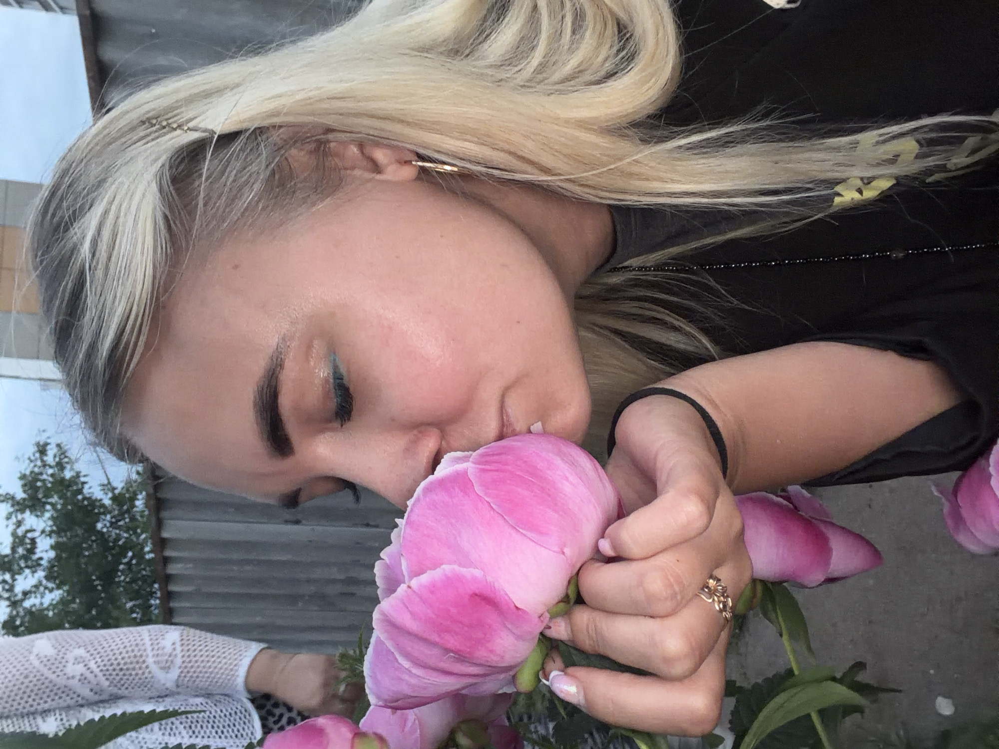 A young blonde woman with light skin and blue eyeliner is holding a large pink peony to her face and sniffing it. She is wearing a black t-shirt and a gold hoop earring. Her right hand, with manicured nails, is gently cupping the flower. On her ring finger, she wears a delicate gold ring. A black beaded necklace hangs down her chest. In the background, a grey corrugated metal fence and trees are visible under a cloudy sky. A portion of another person's white lace sleeve is visible on the far left of the image. The setting appears to be an outdoor area, likely a garden or backyard, with a concrete-like ground. This image was taken in Chaikovsky, Russia.