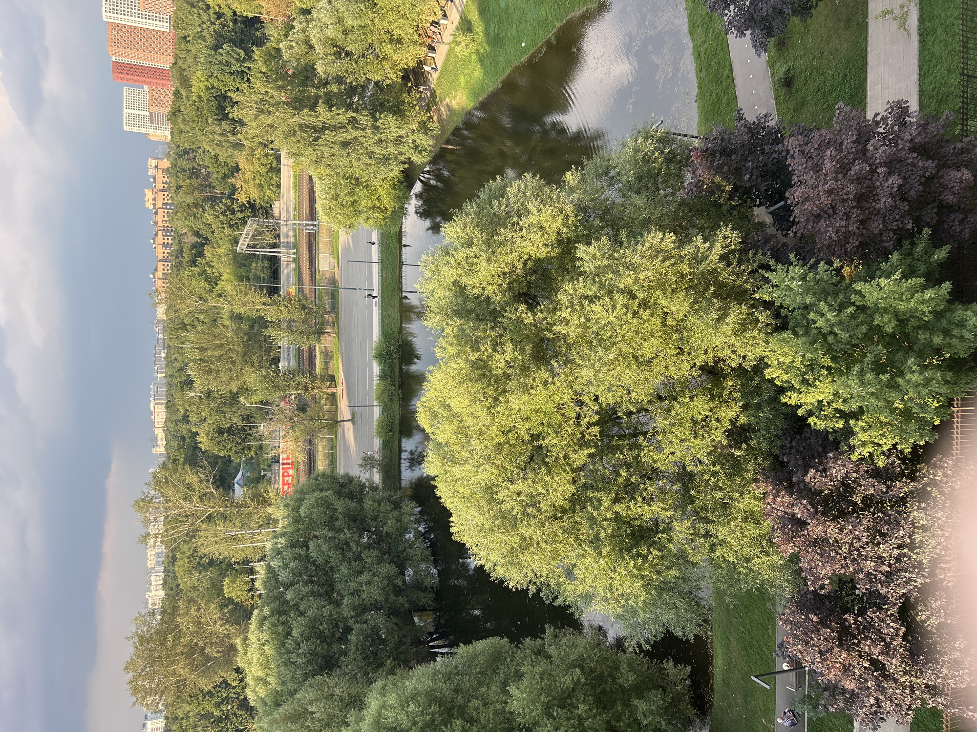 The image shows an aerial view of a park in Lyubertsy, Russia. A dark body of water, likely a pond or canal, meanders through the center of the scene, reflecting the overcast sky and surrounding trees. Lush green foliage dominates the landscape, with various types of trees, some with lighter green leaves, others with darker, more reddish-purple leaves. A paved pathway runs along the edge of the water, with several people visible walking or sitting on benches. In the background, a road with a few pedestrians can be seen, and beyond that, railway tracks and overhead power lines. Further in the distance, a cityscape unfolds with various buildings, including apartment complexes and some with distinctive red accents. The overall impression is of an urban green space, likely a recreational area within the city.