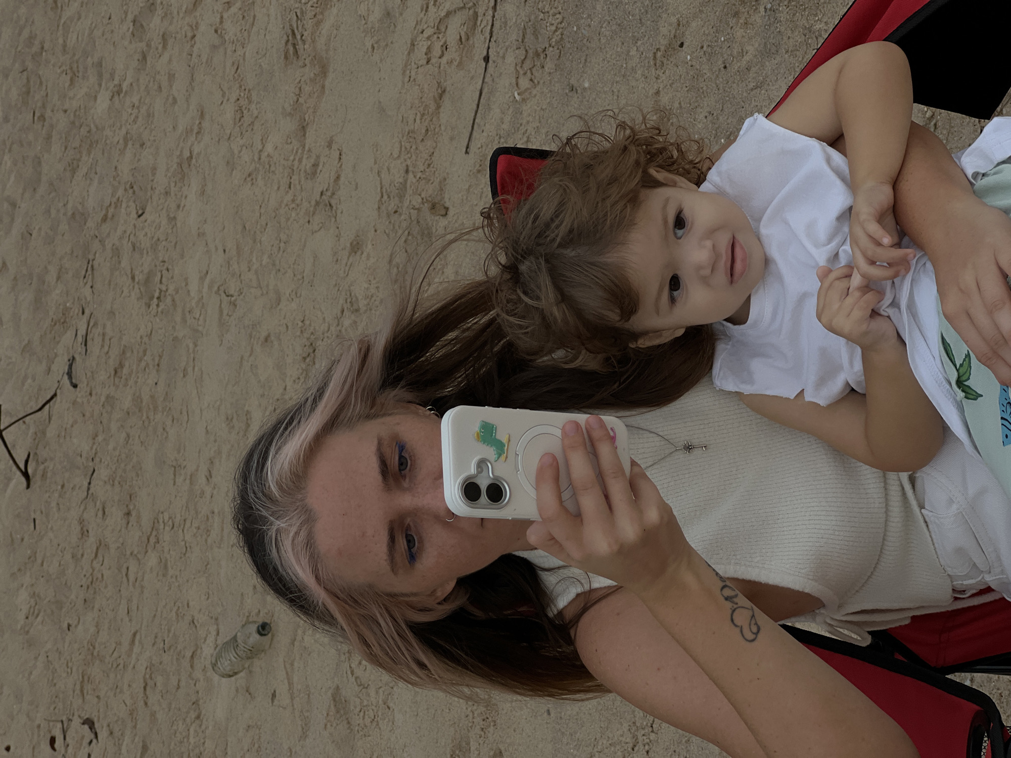 A woman is sitting in a red camping chair on a sandy beach, holding a cell phone in her left hand. A young child is sitting on her lap, with the woman's right arm around the child. The woman has long, brown hair with pink highlights and is wearing a white sleeveless top. She has a tattoo on her left forearm. The child has curly brown hair and is wearing a white t-shirt. In the background, the sand is littered with a few pieces of debris, including a plastic bottle. The image appears to have been taken at eye level, with the subjects in the foreground and the beach extending into the background.