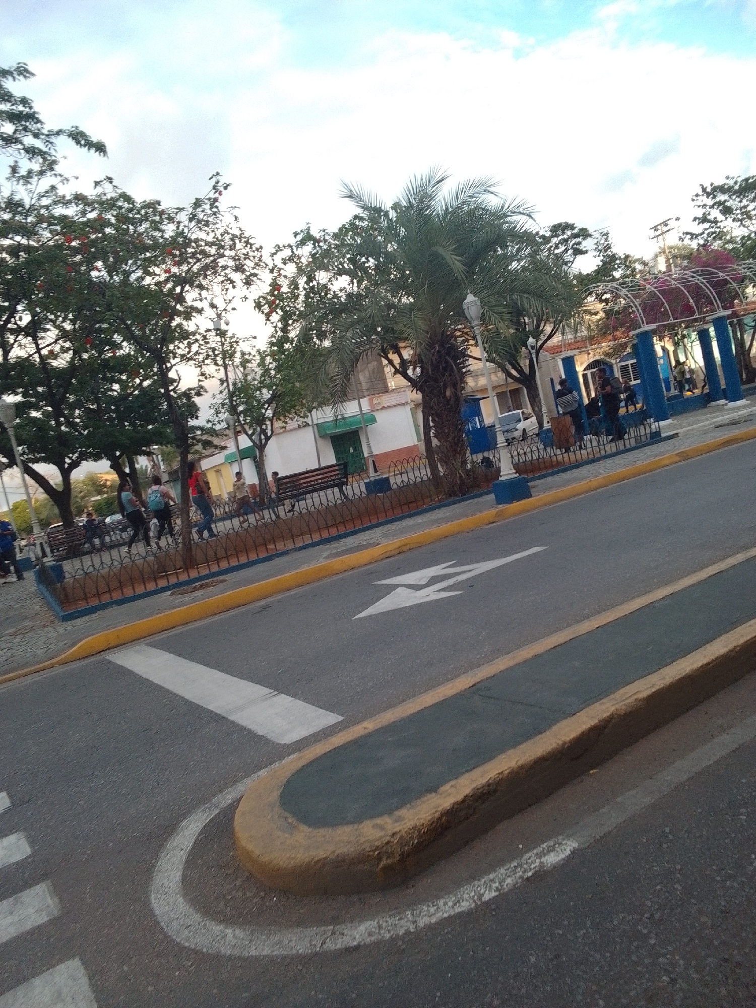 This media file shows a street scene in La Tagua, Venezuela, likely in the late afternoon or early evening given the lighting. The foreground features a paved road with a pedestrian crossing marked by white stripes and a yellow-painted curb. A curved, raised island separates the road from a sidewalk area.
Beyond the road, a park-like area is visible, bordered by a decorative metal fence with blue posts. Several trees, including a palm tree and trees with red blossoms, are present. Benches are situated within this area, and a white building with green trim and a green door is visible in the background, possibly a public building or shop. A blue and white arched structure, perhaps a gazebo or decorative entrance, is also part of the park.
Several people are scattered throughout the park area. Some are walking, while others appear to be sitting or standing. One woman is wearing a backpack. The atmosphere appears relaxed and casual, suggesting a normal day in a public space. The sky is partly cloudy with a light blue hue. There are no discernible signs with visible text in the image.