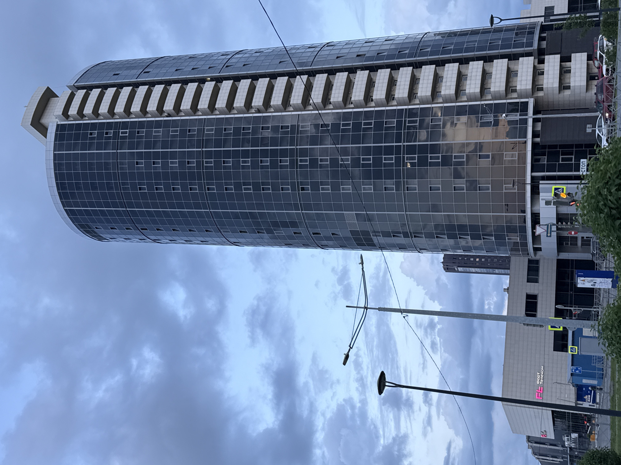 An outdoor, slightly elevated, medium shot captures a modern, cylindrical glass-faced skyscraper in Perm, Russia, against a cloudy, twilight sky. The building's facade is a grid of dark glass panels with vertical white supports, and a series of protruding white balconies ascend the right side of the tower. To the left of the skyscraper, a lower, rectangular building with a textured light-colored facade is visible, featuring a prominent pink and white sign that reads "ФОРТ Телеком". Streetlights are positioned in the foreground, and several cars are parked or driving on a street running in front of the buildings. A traffic light displaying yellow is visible. The overall atmosphere is urban and contemporary, with the overcast sky suggesting cooler weather or evening hours.