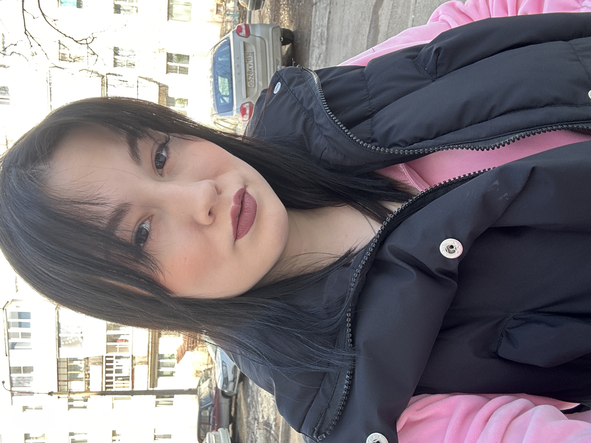 A medium-close, tilted portrait shows a young woman with dark hair and fair skin against an urban backdrop, likely in Chaikovsky, Russia given the car's license plate. She wears a black puffer jacket over a pink hoodie. Her lips are a deep rose-brown. Behind her are apartment buildings, leafless tree branches, and parked cars including a light-colored car with Russian license plate O528KY159. The lighting is soft and diffuse, suggesting either an overcast day or a position in partial shade.