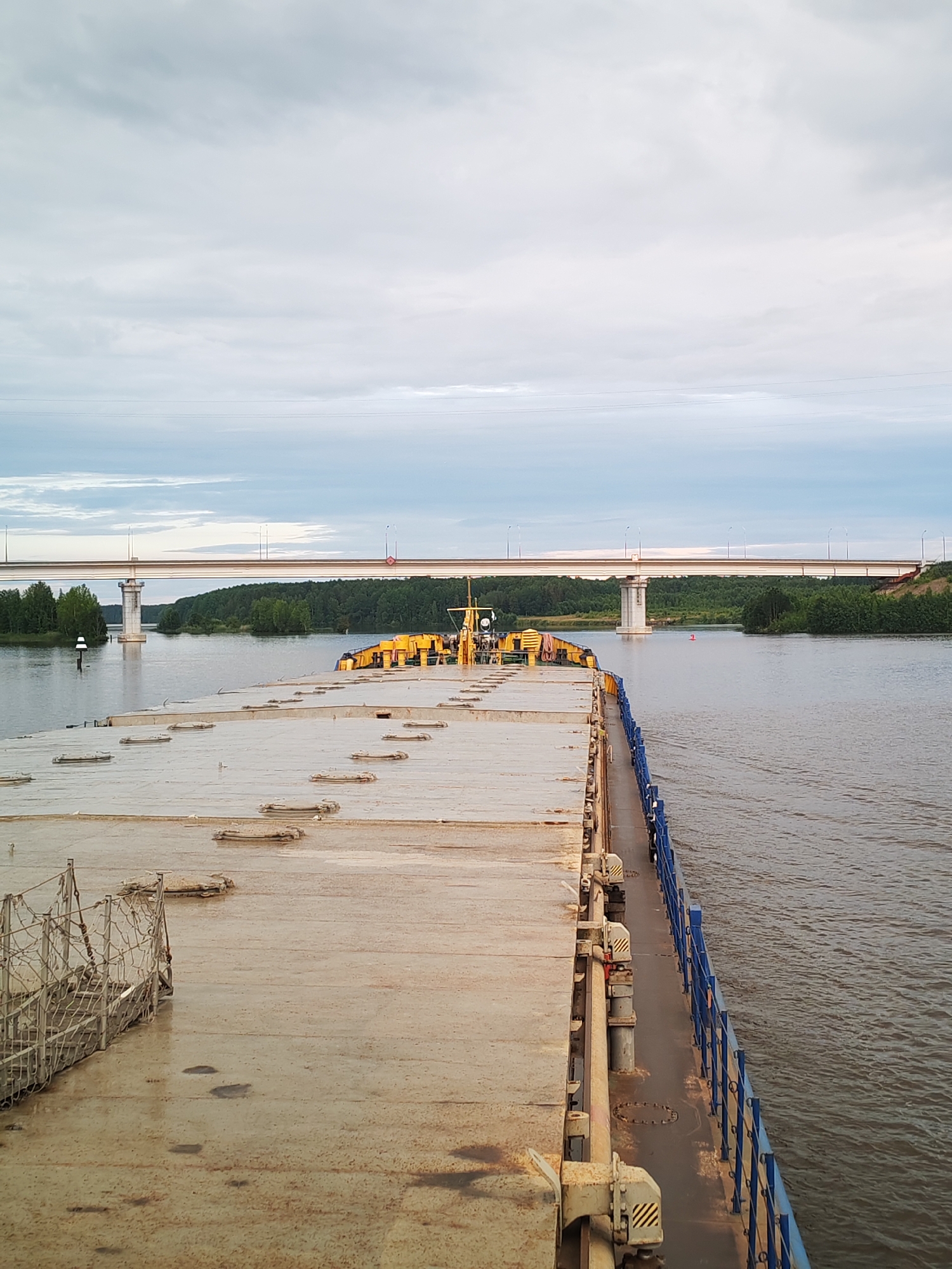 The image shows a perspective from the deck of a barge moving on a river, heading towards a bridge. The barge's deck is a large, flat, weathered surface with several rectangular hatches. A blue railing runs along the right side of the barge. In the distance, a wide river is visible, bordered by a dense green forest on either side. A large, modern bridge with multiple lamp posts spans the river. The sky is overcast with light gray clouds. The scene appears to be in Sheksna, Russia, given the presence of a bridge over a river, which is a common feature of the region. There are no people visible in the image. The barge is the primary subject, with the bridge and the river as background elements. The overall atmosphere is calm and serene, typical of a waterway in a rural or semi-rural setting.