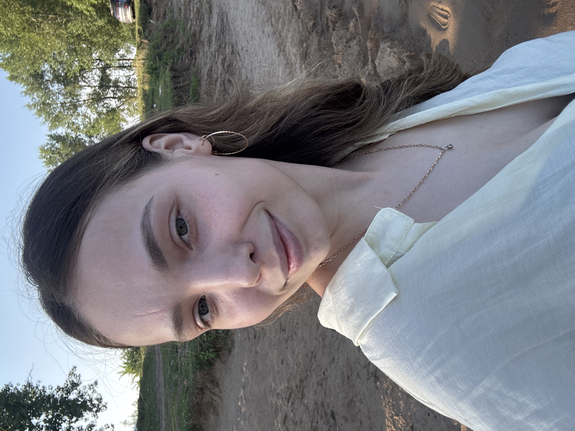 A young woman with long, dark brown hair and fair skin is posing for a selfie on a sandy embankment. She is wearing a light yellow collared shirt and a delicate necklace with a small pendant. Her right ear is adorned with a gold hoop earring. The background consists of a clear blue sky, a line of trees, and a sandy beach with footprints. In the distance, a dark-colored car is visible on a road. The lighting suggests it is either early morning or late afternoon. The overall scene appears to be a natural outdoor setting, possibly near a body of water.