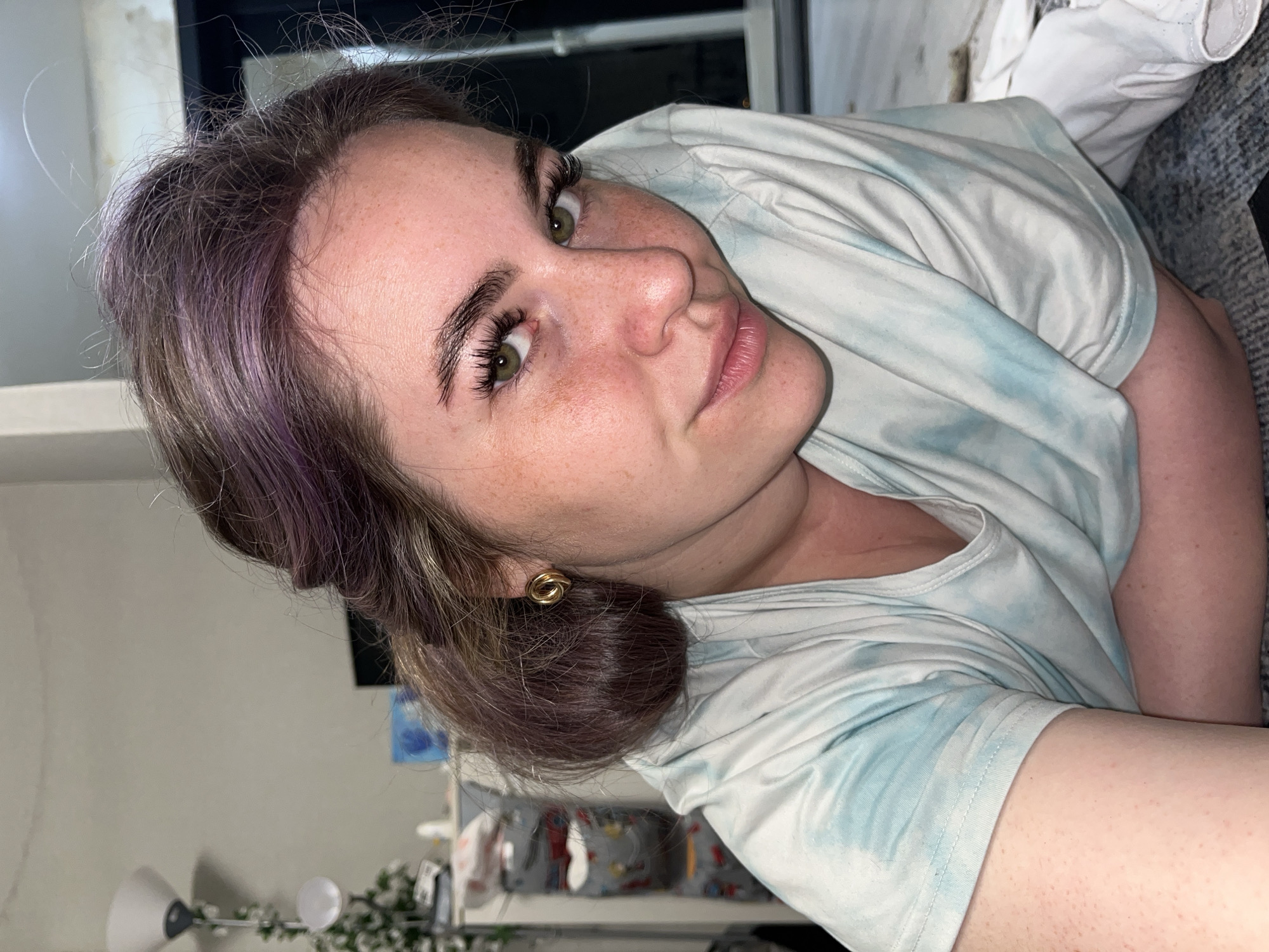 A woman with fair skin, freckles, green eyes, and dark brown hair with purple highlights is looking at the camera with a slight smile. Her hair is styled in a bun, and she is wearing gold earrings. She is wearing a light blue and white tie-dye t-shirt. The background appears to be an interior setting with a lamp, a window, and other household items. The location in the city of Chaikovsky, Russia is unknown.